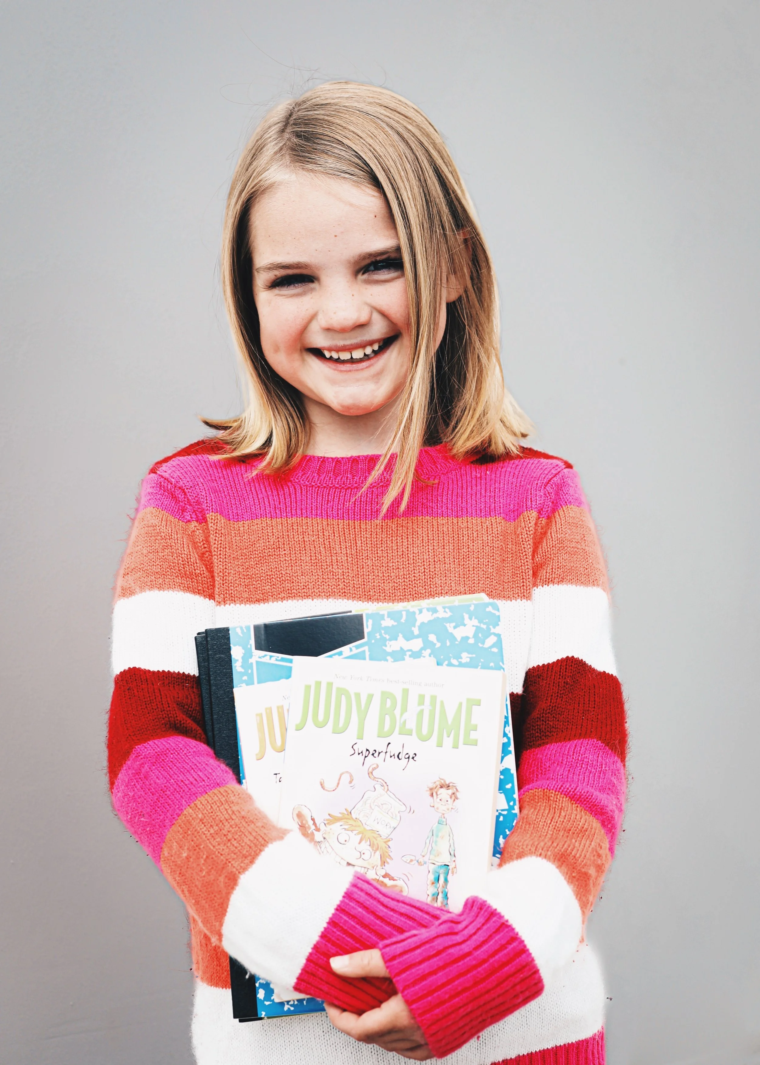 A smiling young girl with shoulder-length blonde hair holding multiple Judy Blume books, wearing a colorful striped sweater.
