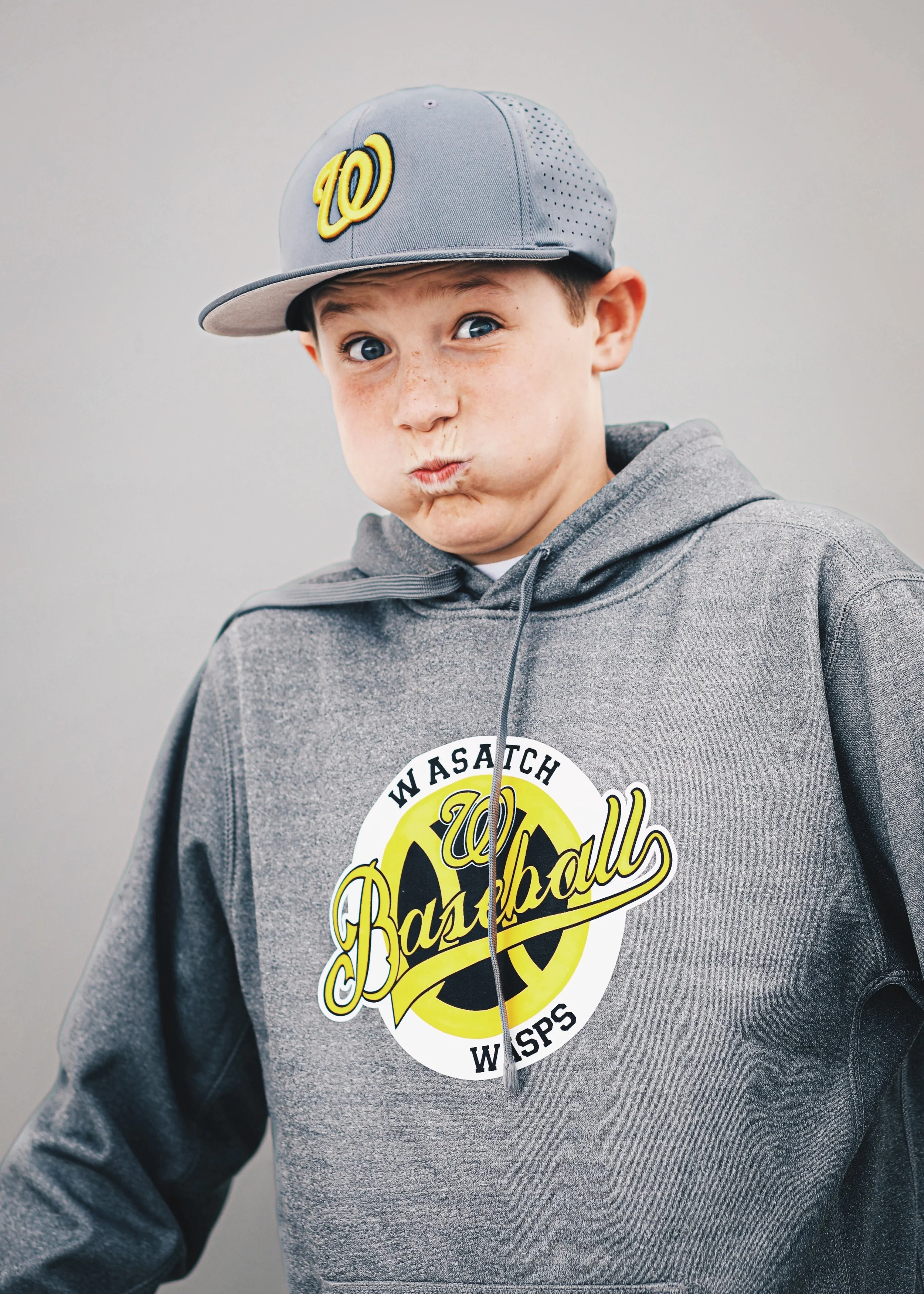 A young boy wearing a gray Washington Baseball sweatshirt and a gray baseball cap with a yellow 'W' logo, making a playful pouty face.