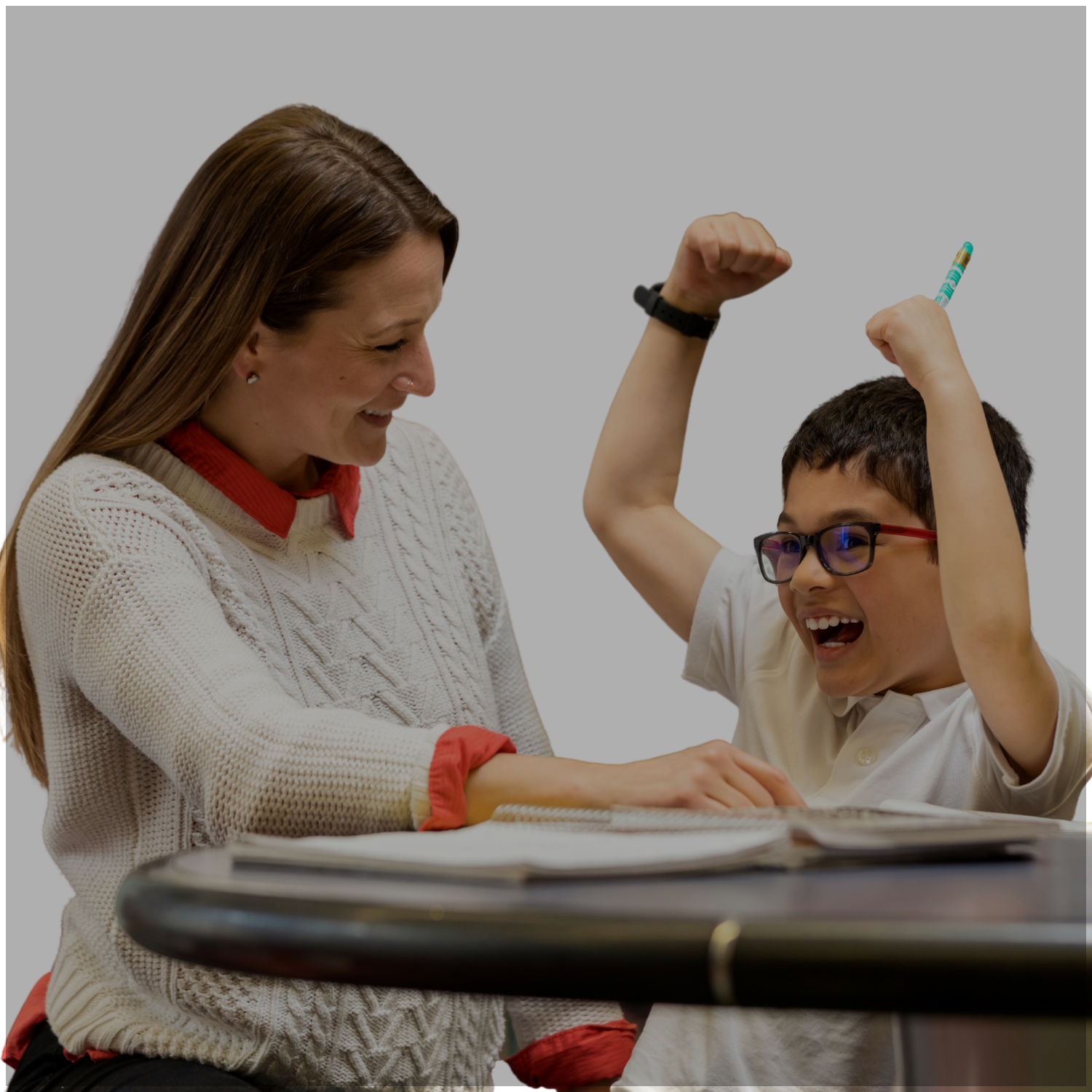A smiling woman and a boy with glasses celebrating together at a table, the boy raising his arms in excitement, with an open book on the table.