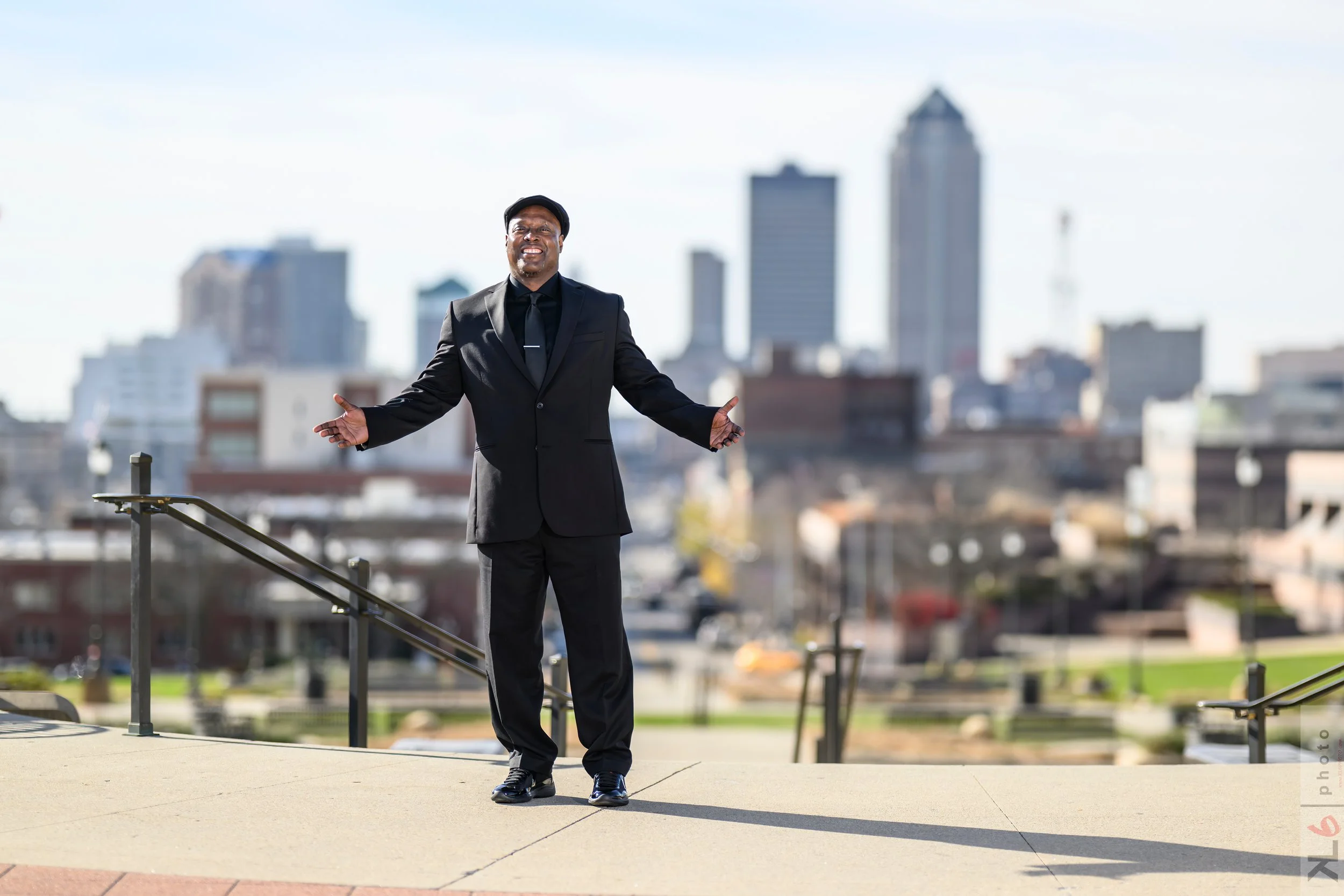 Man in a black suit standing outdoors with arms open, smiling, with a city skyline in the background.