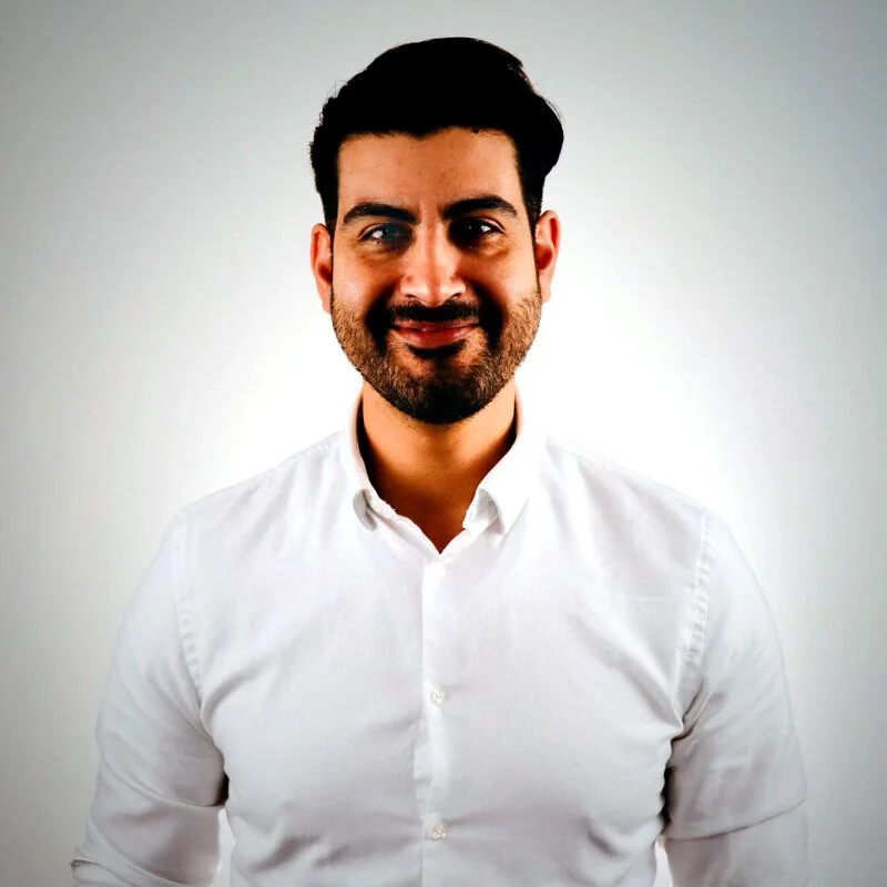 A man with dark hair and a beard, wearing a white button-up shirt, standing against a plain light gray background.