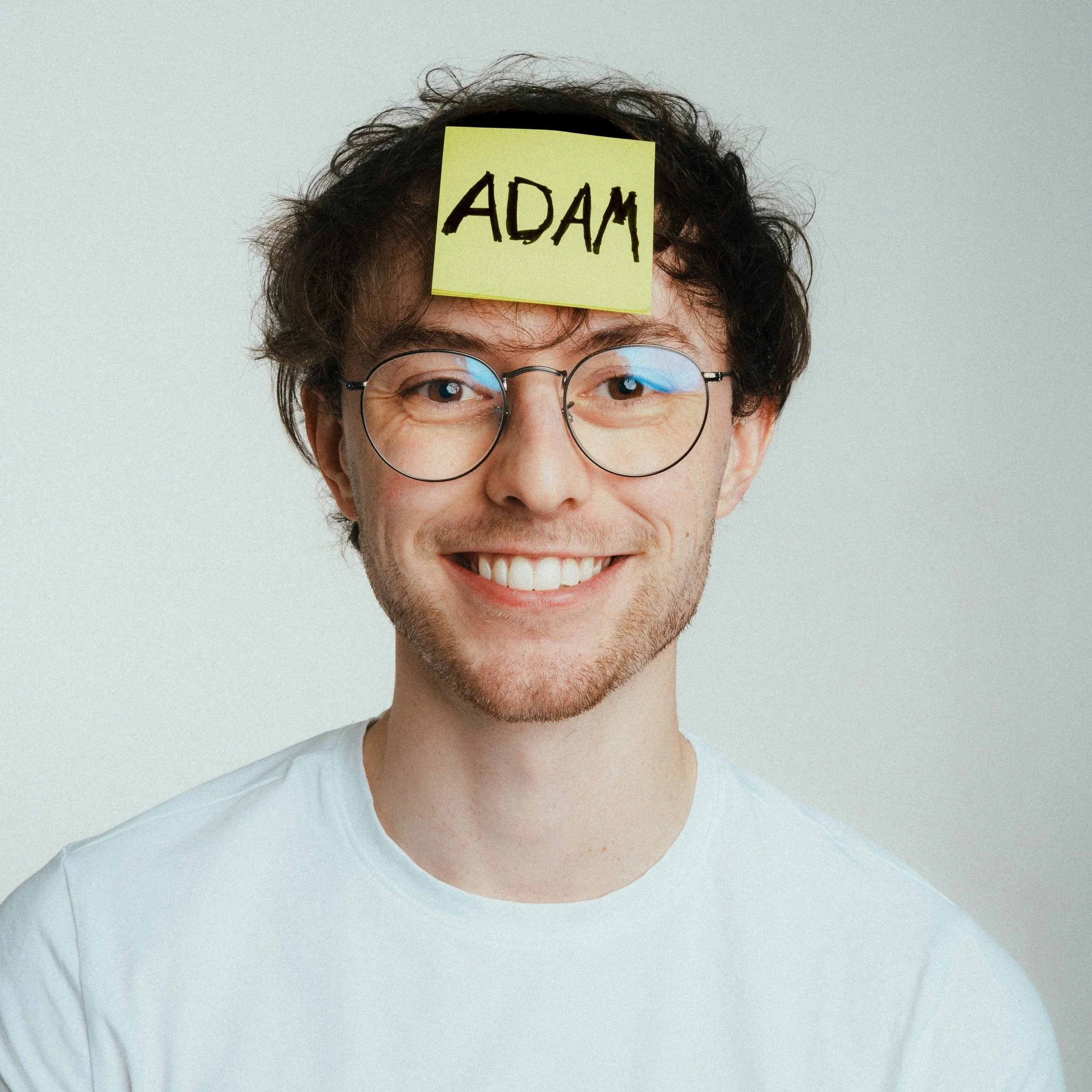 Person with curly hair and glasses smiling, with a yellow sticky note on forehead that reads 'ADAM'.
