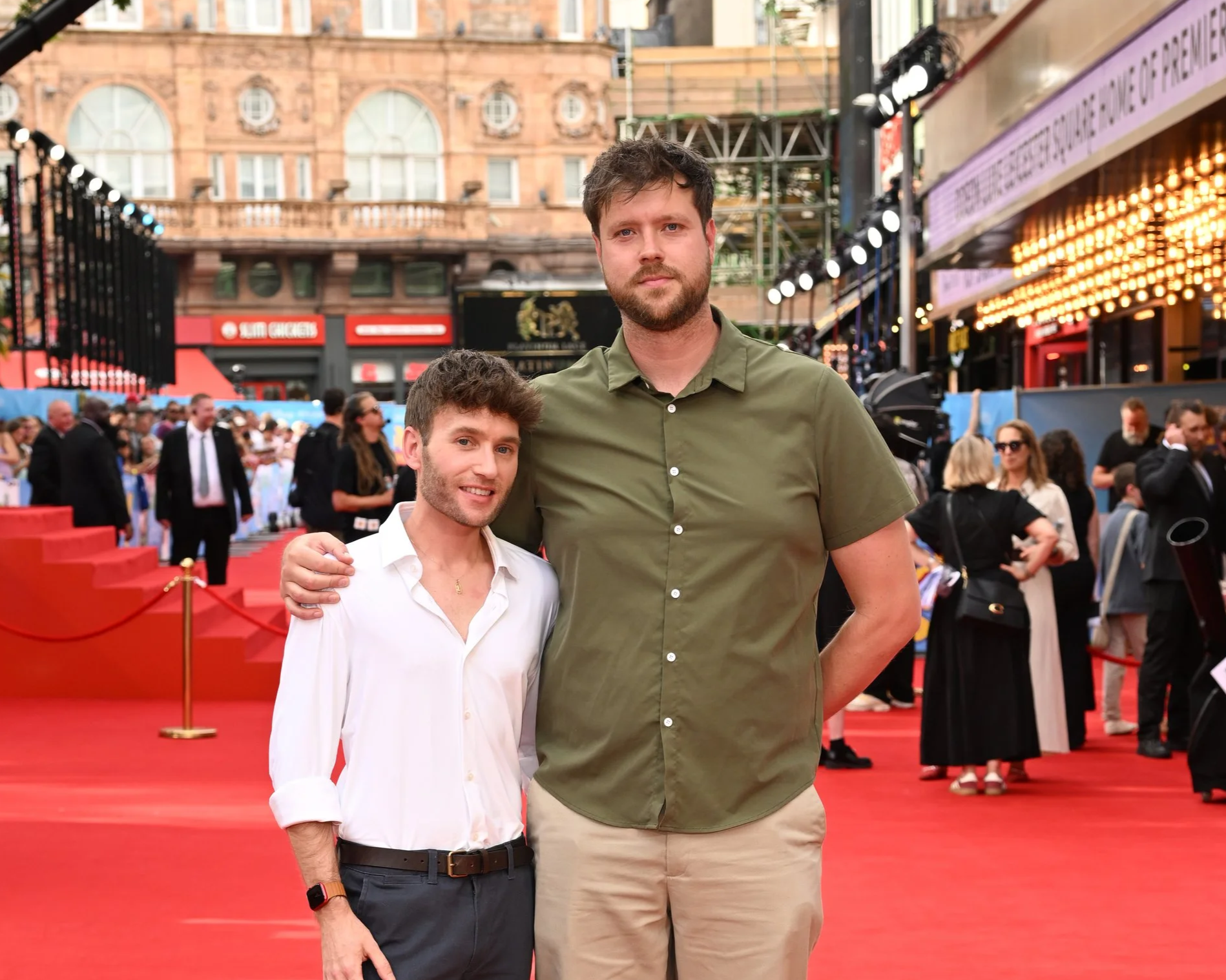 Two men standing side by side on a red carpet at a formal event, with a crowd in the background. One man is wearing a white shirt and the other a green shirt. The scene is set outside with historic buildings and some lighting equipment visible.