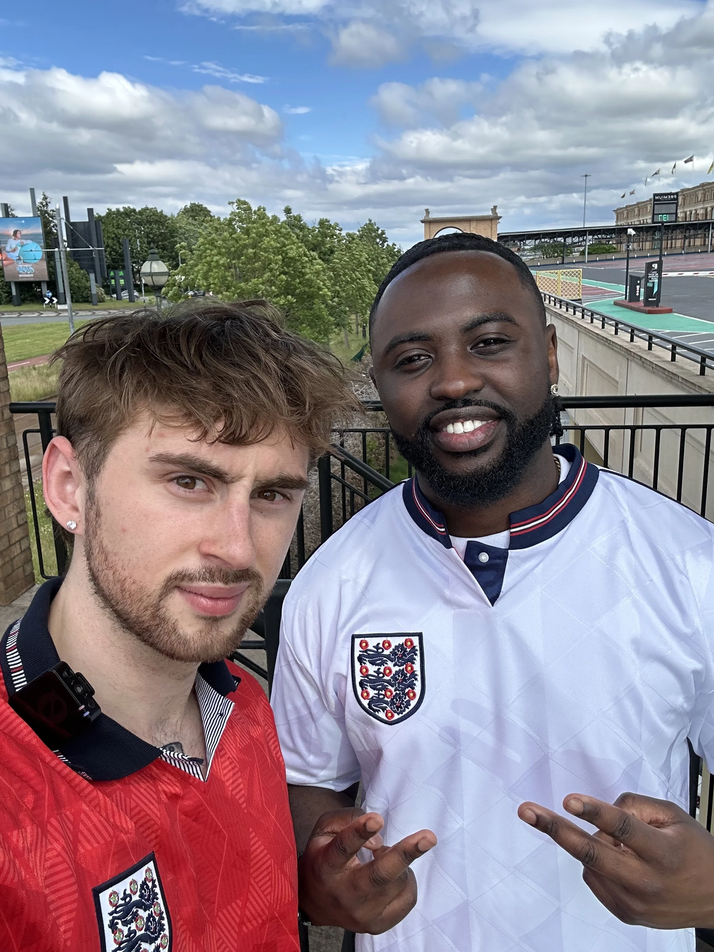 Two men taking a selfie outdoors with a cloudy sky, trees, and buildings in the background. One man has light hair and a beard, wearing a red shirt, and the other is black with a beard, wearing a white football jersey with an England crest.