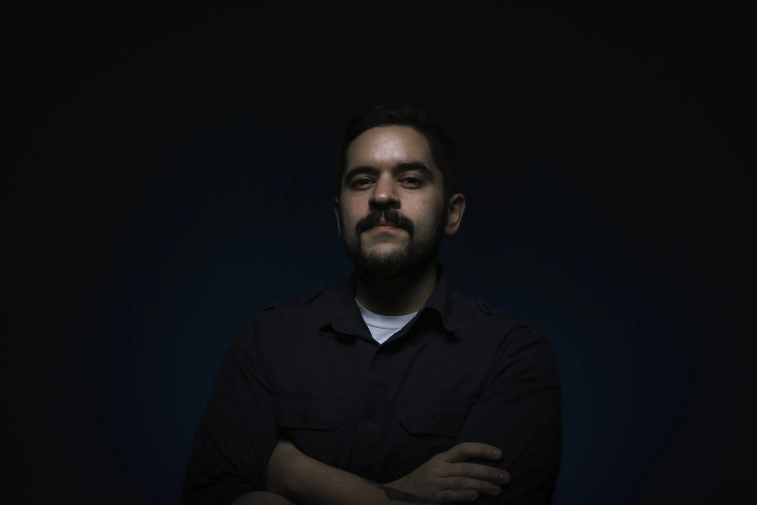 A man with dark hair and a beard wearing a black shirt advances his arms with his left arm crossing his torso and touching his right arm, against a dark background.