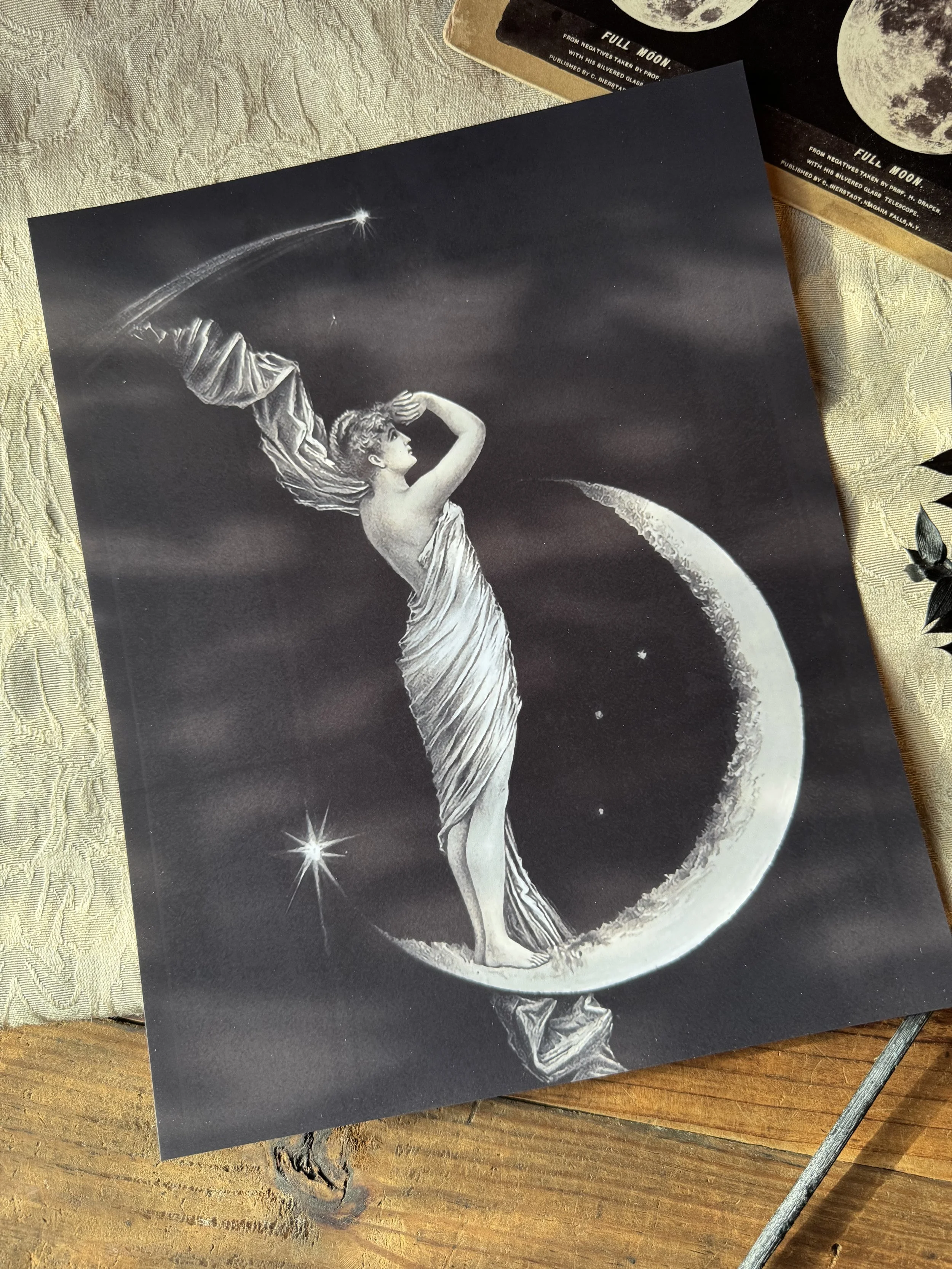 a black and white print with a grecian, cloaked woman standing atop a crescent moon