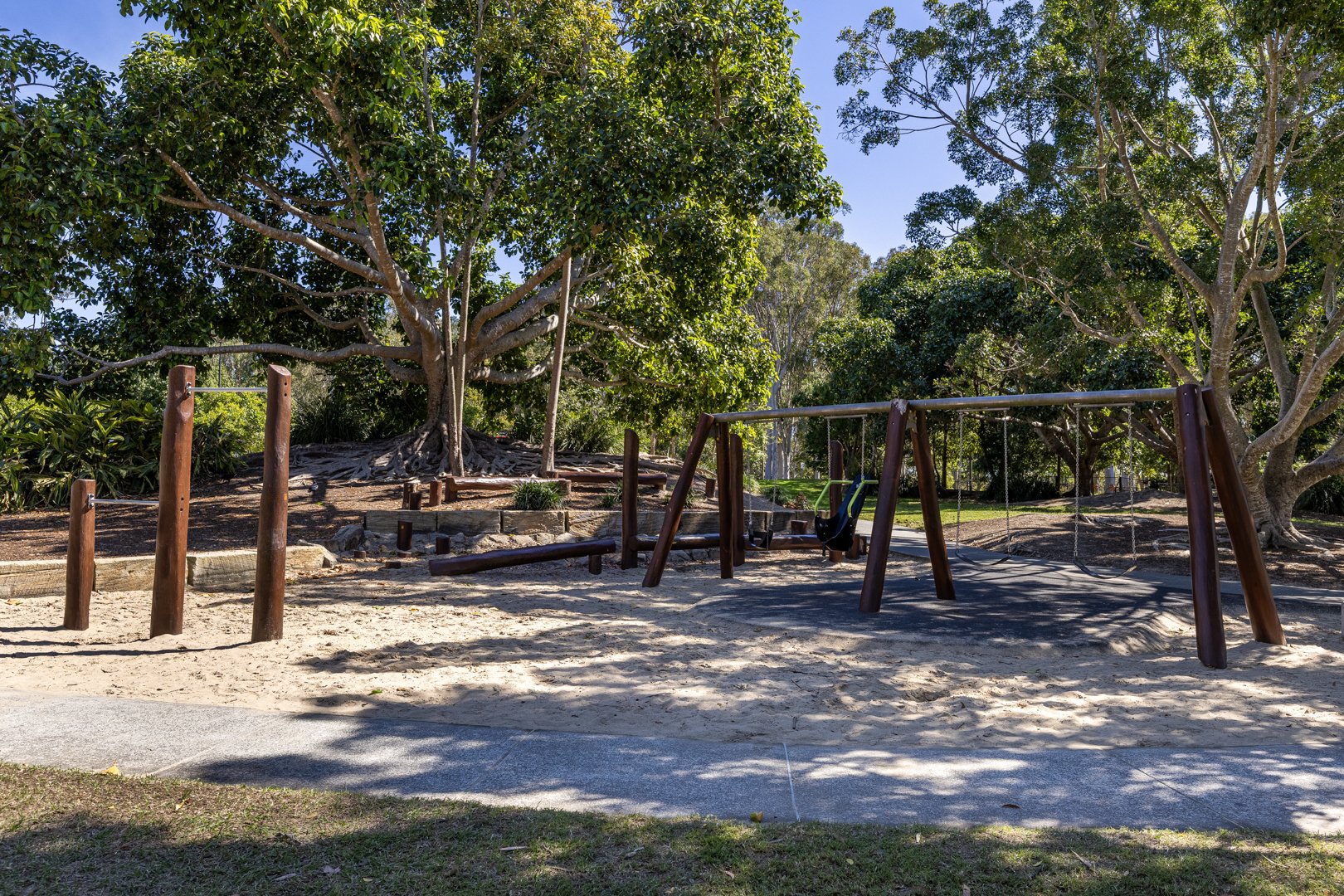 Timber swing set and monkey bars shaded by fig trees at a Logan Garden playground in Logan Central