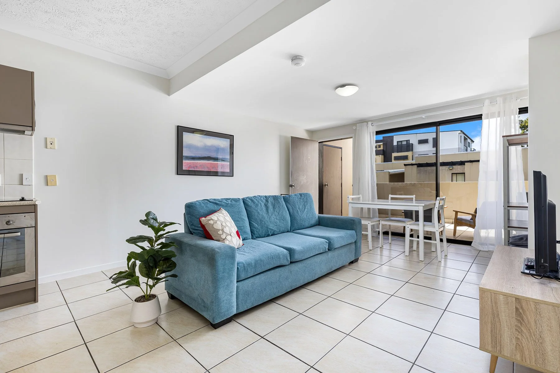 Sun-drenched open-plan living and dining area in a modern Logan townhouse, featuring light tiled floors, a contemporary blue sofa, and seamless transition to a sunny balcony.