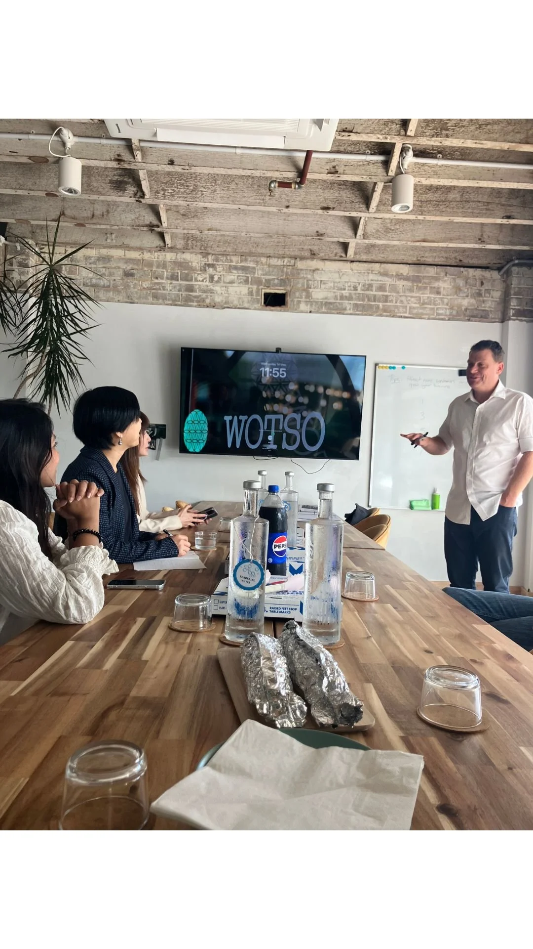 A meeting room with a wooden table, several women sitting on one side, and a man standing near a whiteboard. A large screen on the wall displays the time and the word 'WOTSO' upside down. The table has water bottles, glasses, foil-wrapped food, and a pizza box.