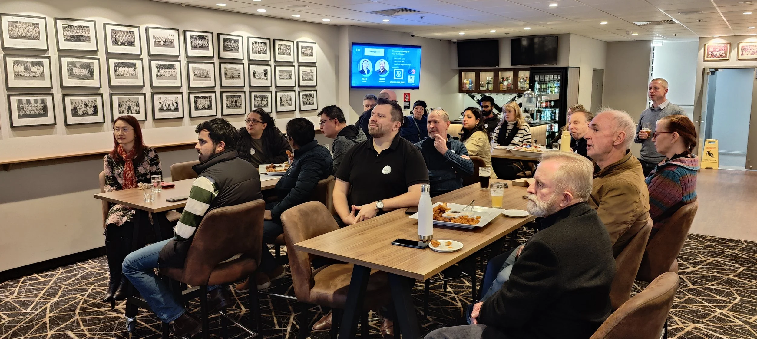 People attending a meetup or presentation in a restaurant or café, sitting at tables with food and drinks, facing a screen, with framed photos on the wall.