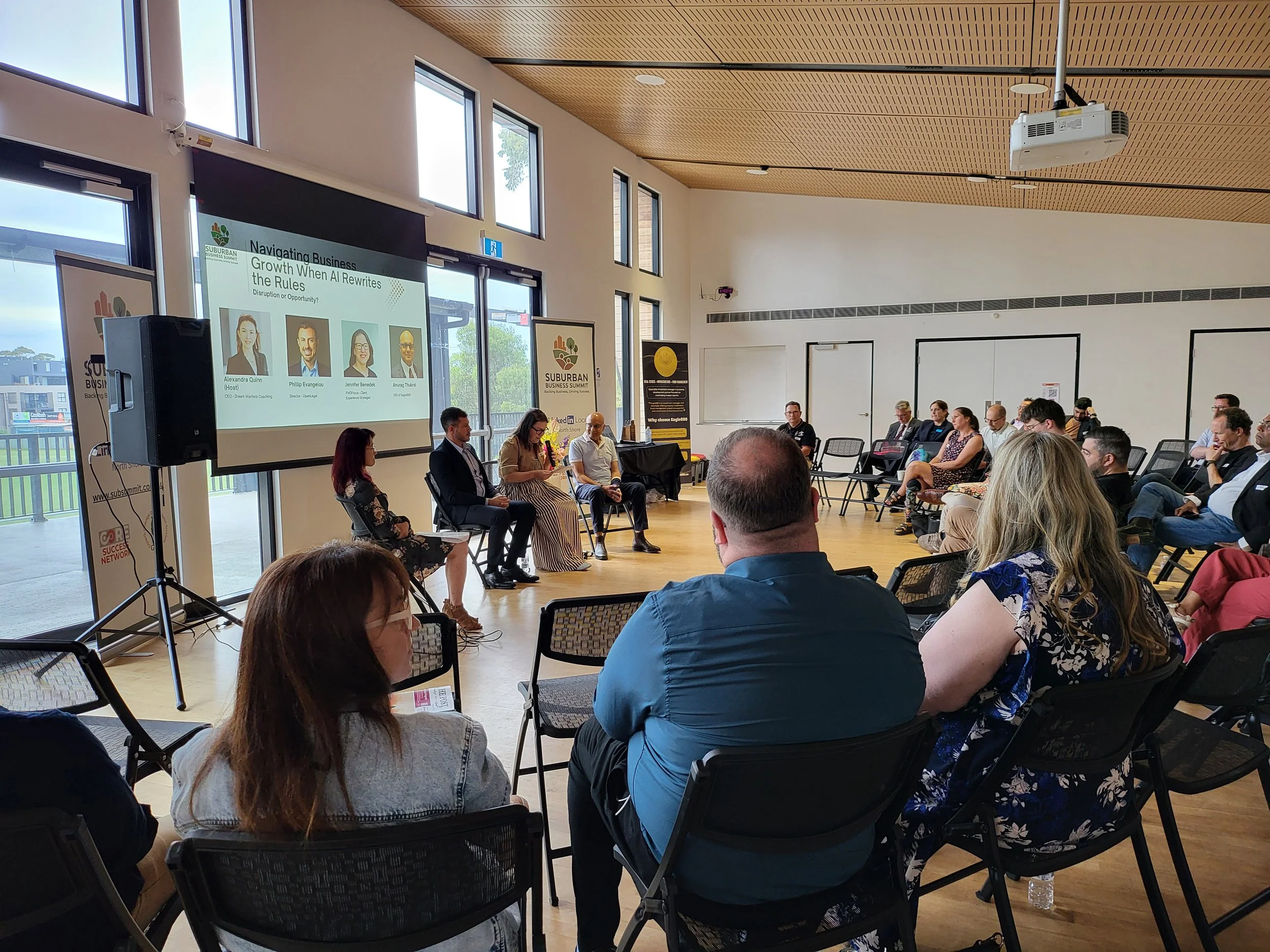 People attending a meetup or presentation in a restaurant or café, sitting at tables with food and drinks, facing a screen, with framed photos on the wall.
