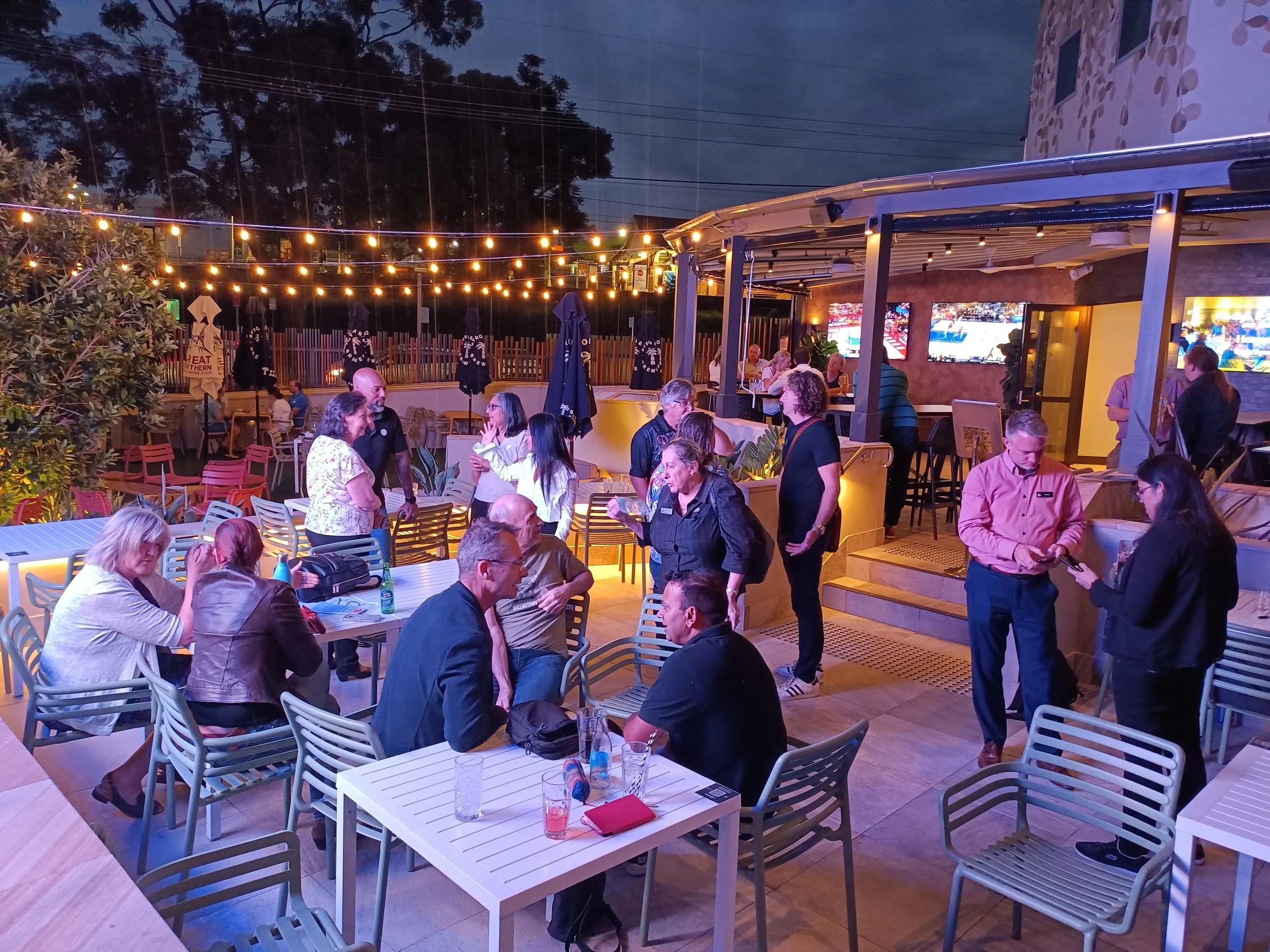 People socialising and dining at an outdoor patio of a restaurant or bar during evening, illuminated by string lights with a covered area, some watching television screens on the wall.