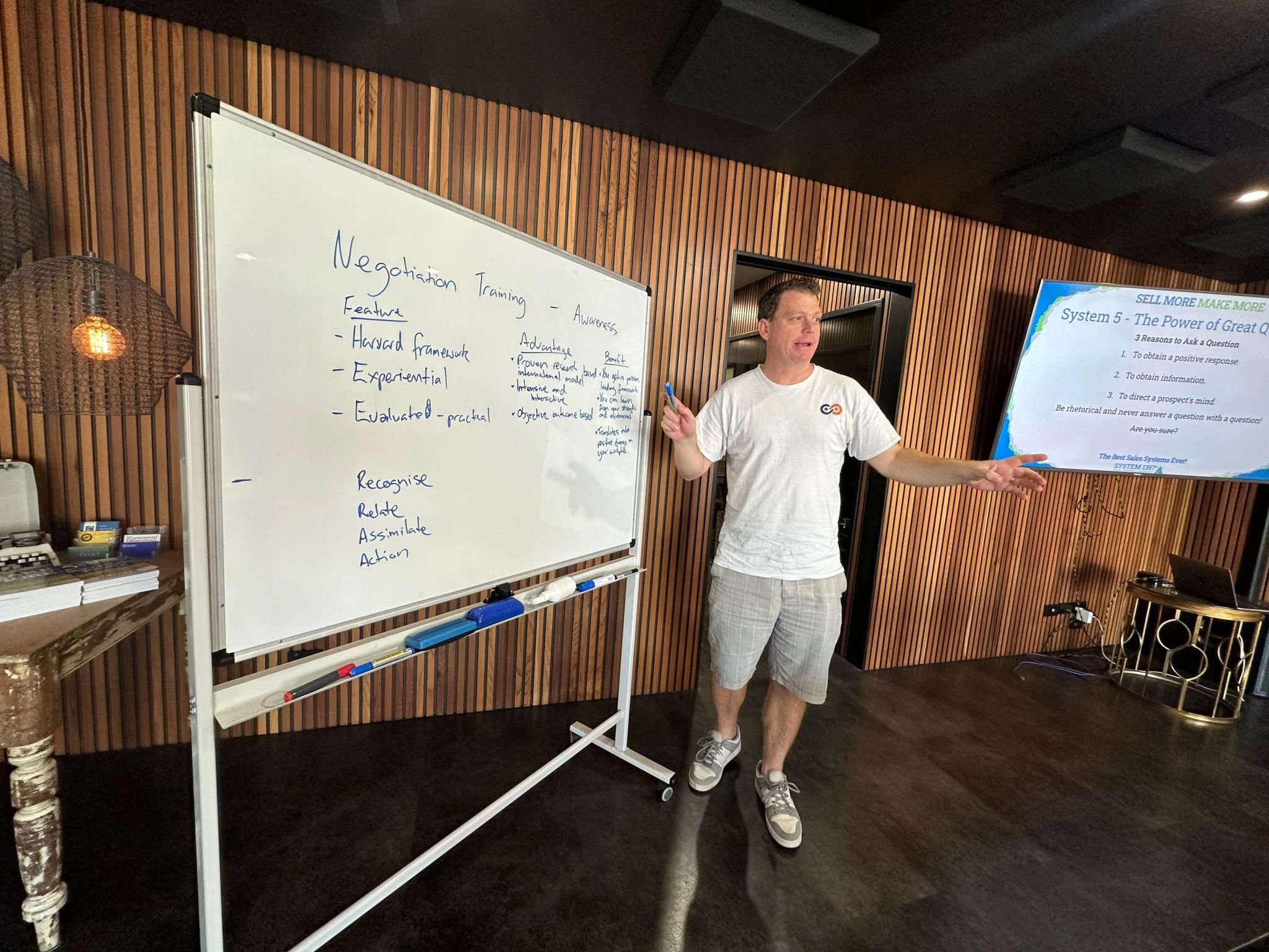 A man giving a presentation in a room with wooden panel walls, standing next to a whiteboard with notes about negotiation training, and a large screen displaying a slide titled 'The Power of Great Q'.