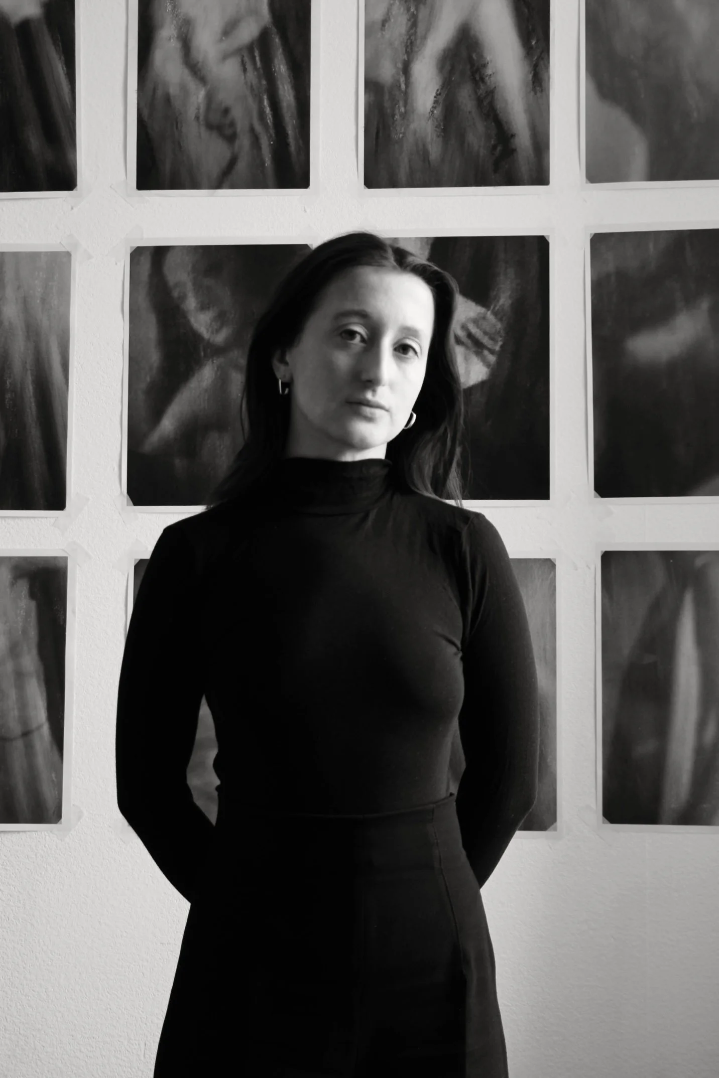 A photograph of Louise standing infant of a series of Motae photographs. She is looking down at the camera, her arms behind her back. She is wearing black polo neck, black trousers, has shoulder length hair, white skin and silver earrings.