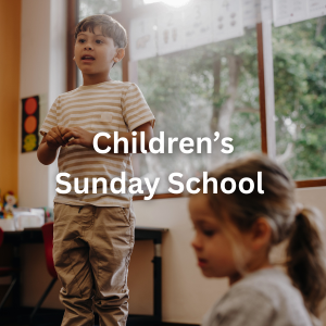 A young boy speaking in front of a classroom during a children's Sunday school class, with a girl sitting nearby and a traffic light decoration on the wall.