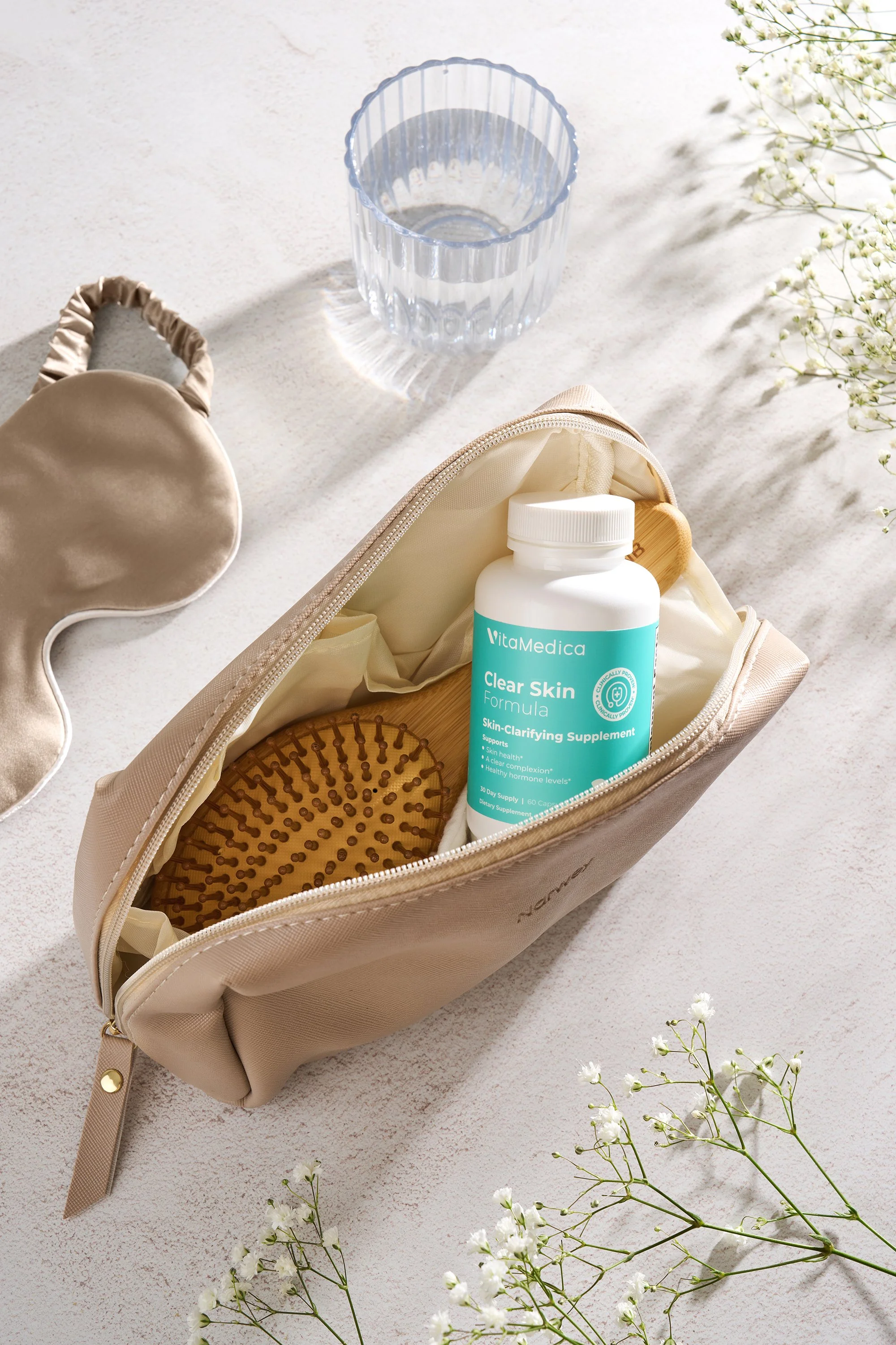 A beige toiletry bag is open with a bottle of VitaMedica Clear Skin supplement and a wooden hairbrush inside. The bag is on a light surface with a clear glass of water, a silk sleep mask, and white flowers nearby.