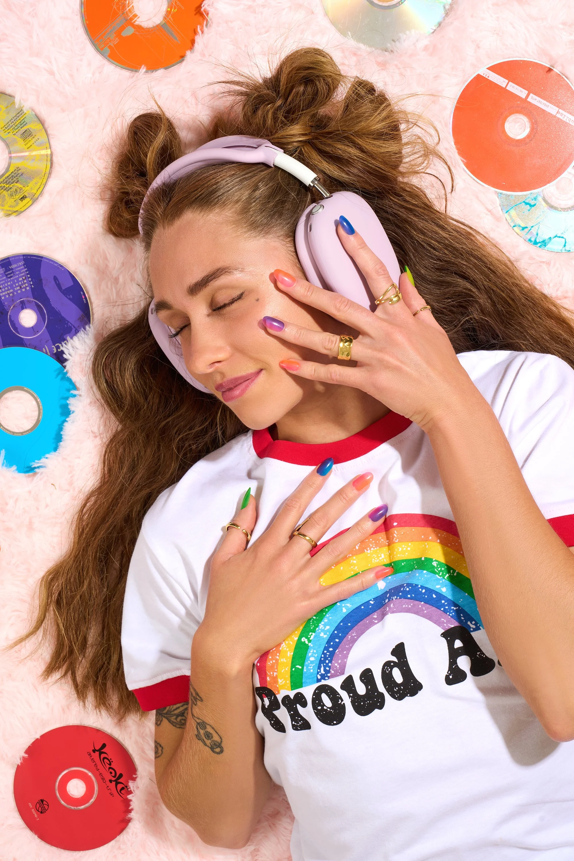 A woman with long, wavy hair lying on a pink furry rug, wearing Kleo Kolor rainbow nails, a white t-shirt with red trim featuring a rainbow and rainbow-colored text, listening to music through pink over-ear headphones, surrounded by music CDs.