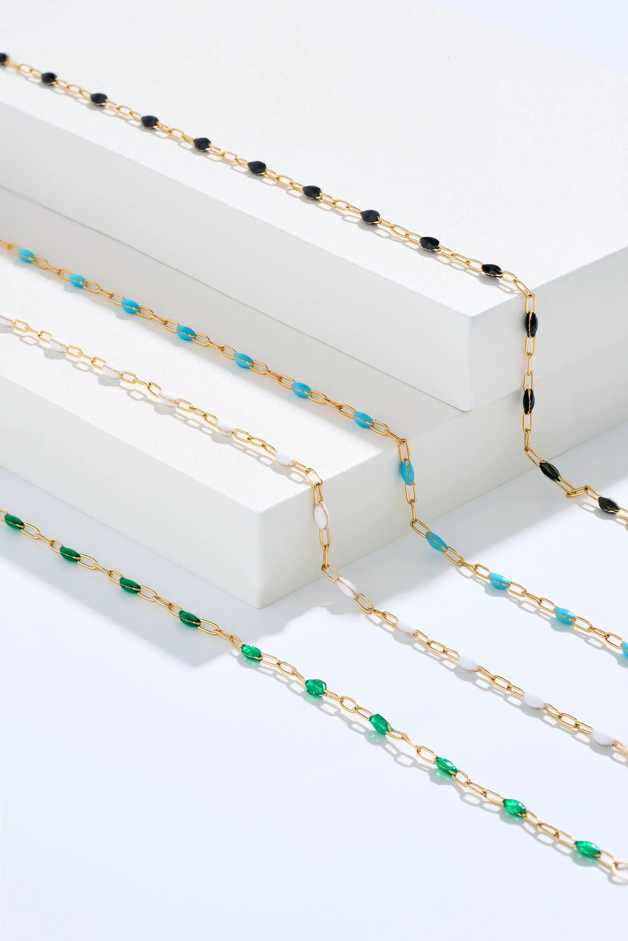 Four colorful beaded necklaces with gold chains are displayed on white platforms.
