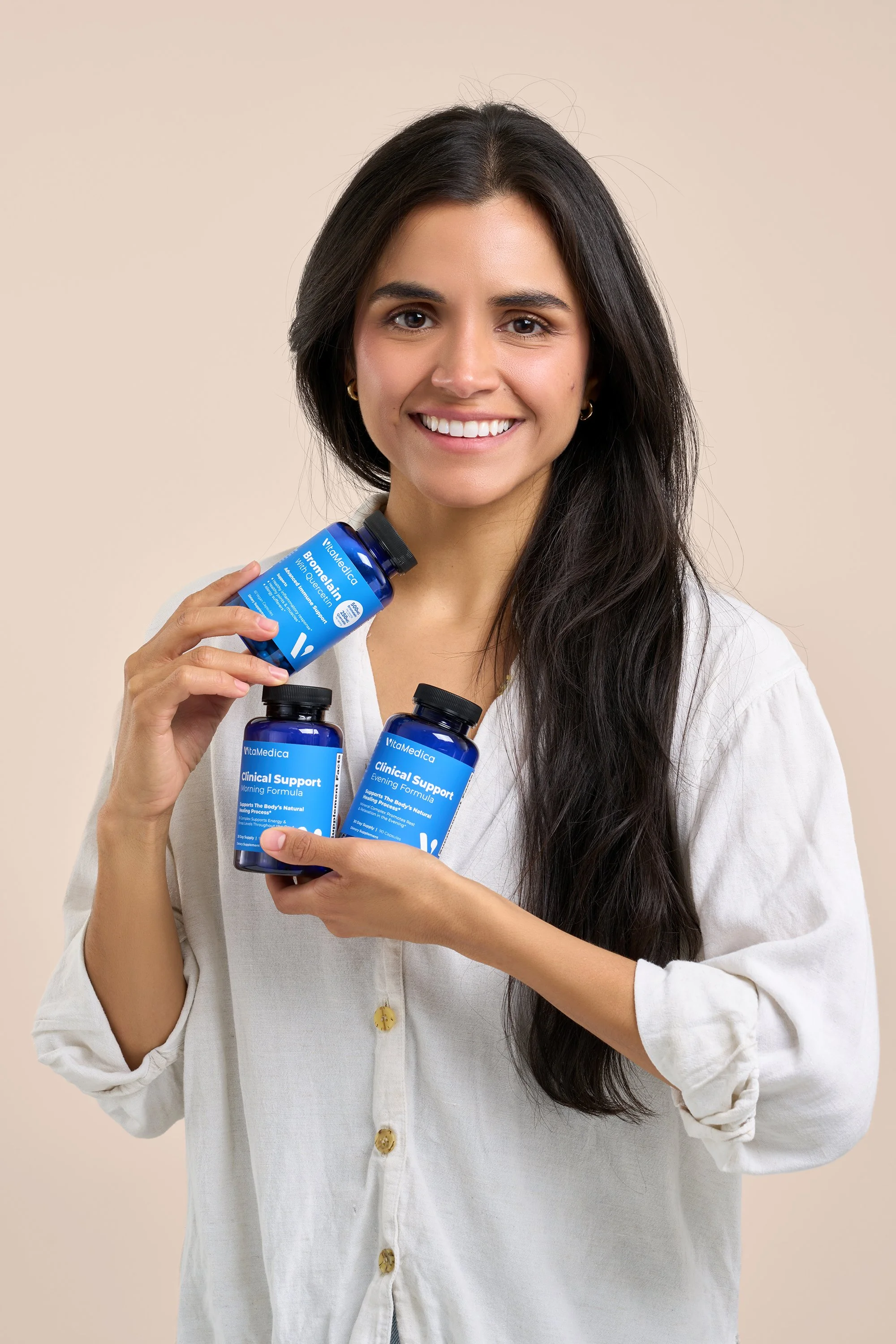 Young woman with long dark hair smiling, holding three blue supplement bottles labeled 'Clinical Support' and 'Bromelain' in front of a beige background.