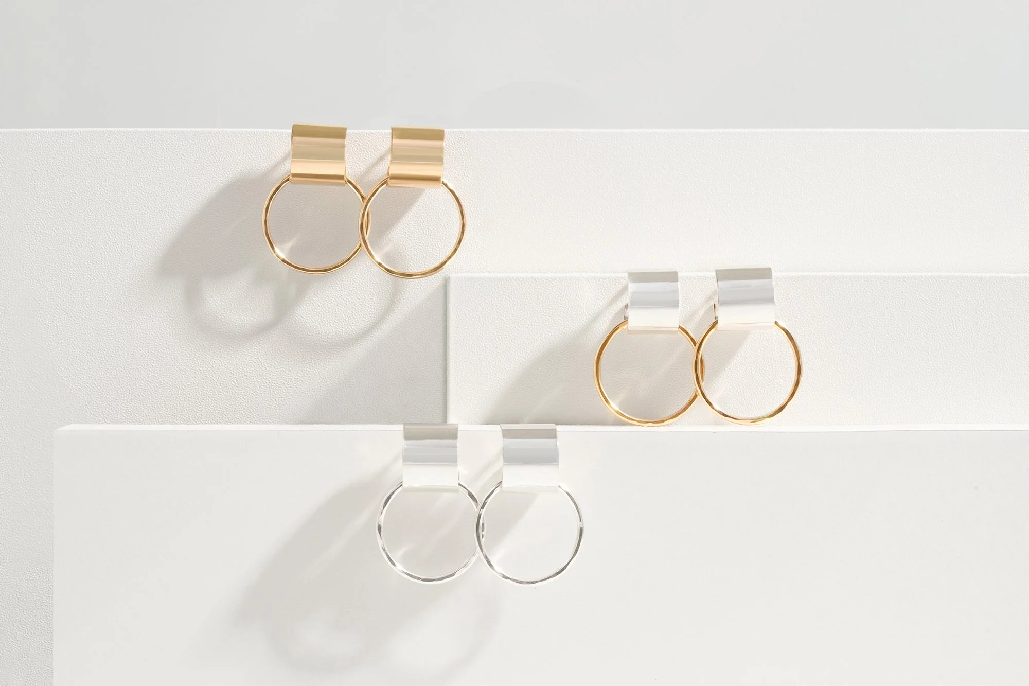 Three pairs of minimalist hoop earrings in gold, silver, and a combination of both, displayed on white platforms with shadows.