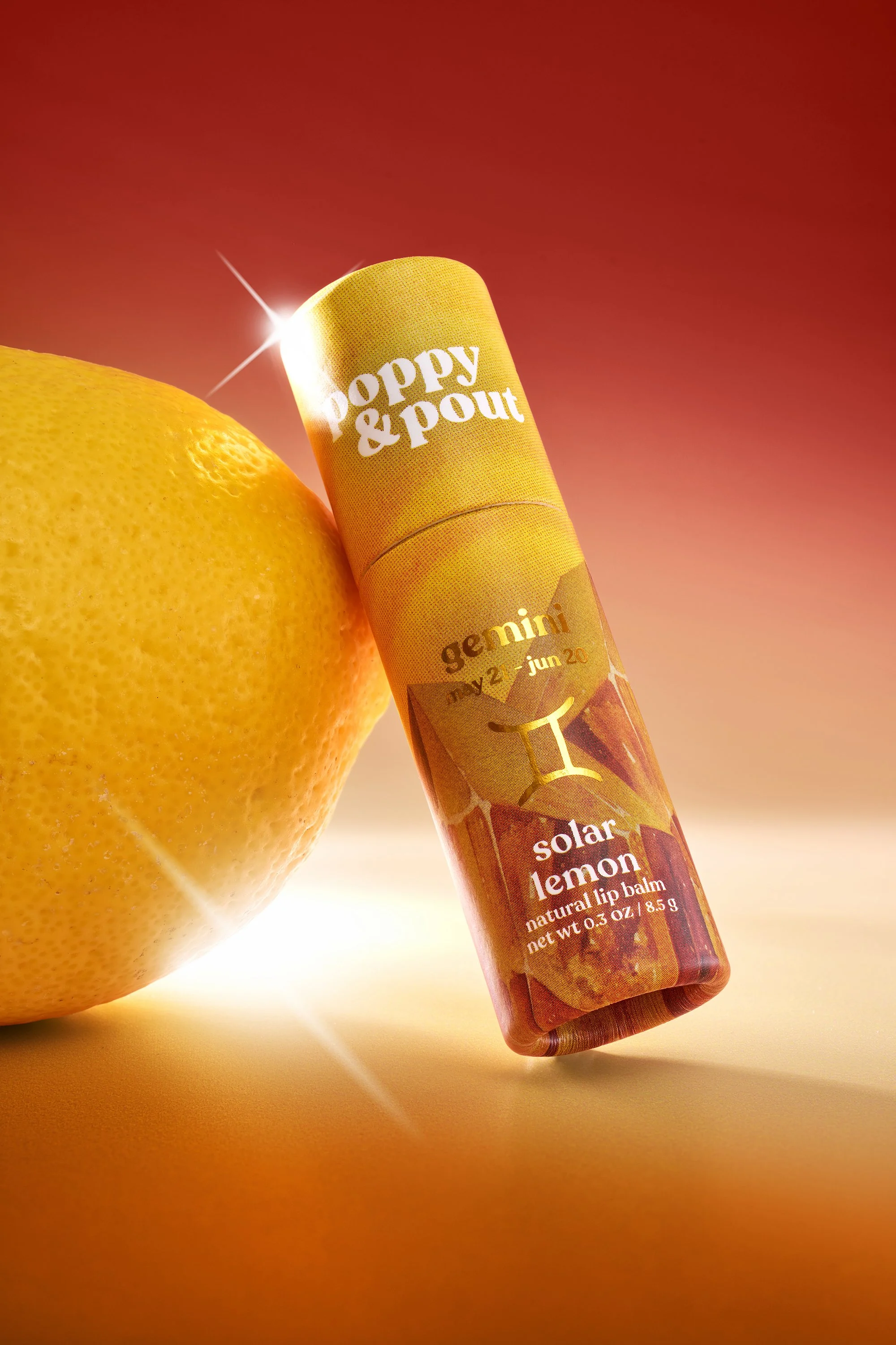 Close-up of a lemon and a tube of Poppy and Pout solar lemon natural lip balm on a warm red gradient background.