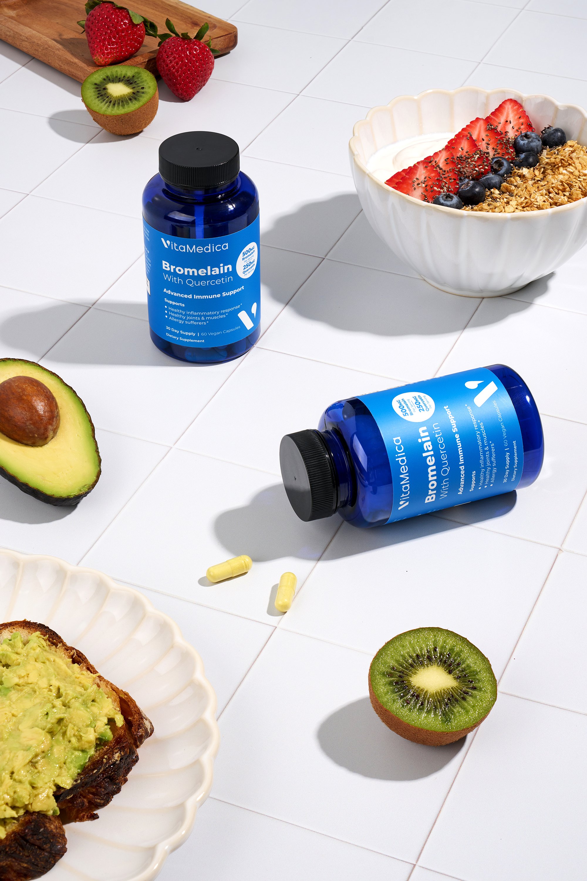 Two blue bottles of VitaMedica Bromelain with Quercetin, some capsules spilled on white tiled surface, with sliced avocado, strawberries, kiwi, and a bowl of yogurt with strawberries, blueberries, granola, and chia seeds.