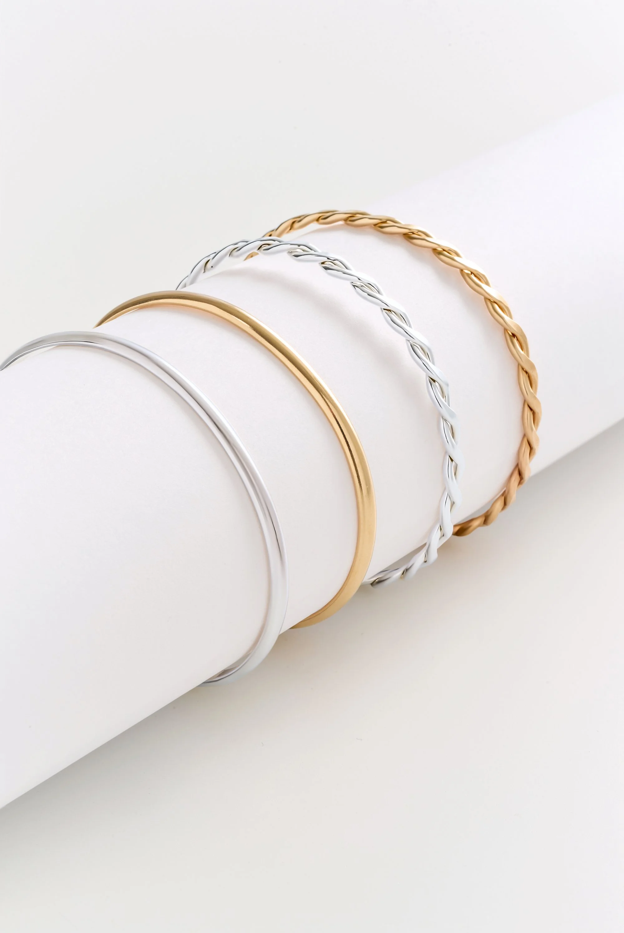 Four metal bangles, in silver, gold, and two braided designs, resting on a white cylindrical display.