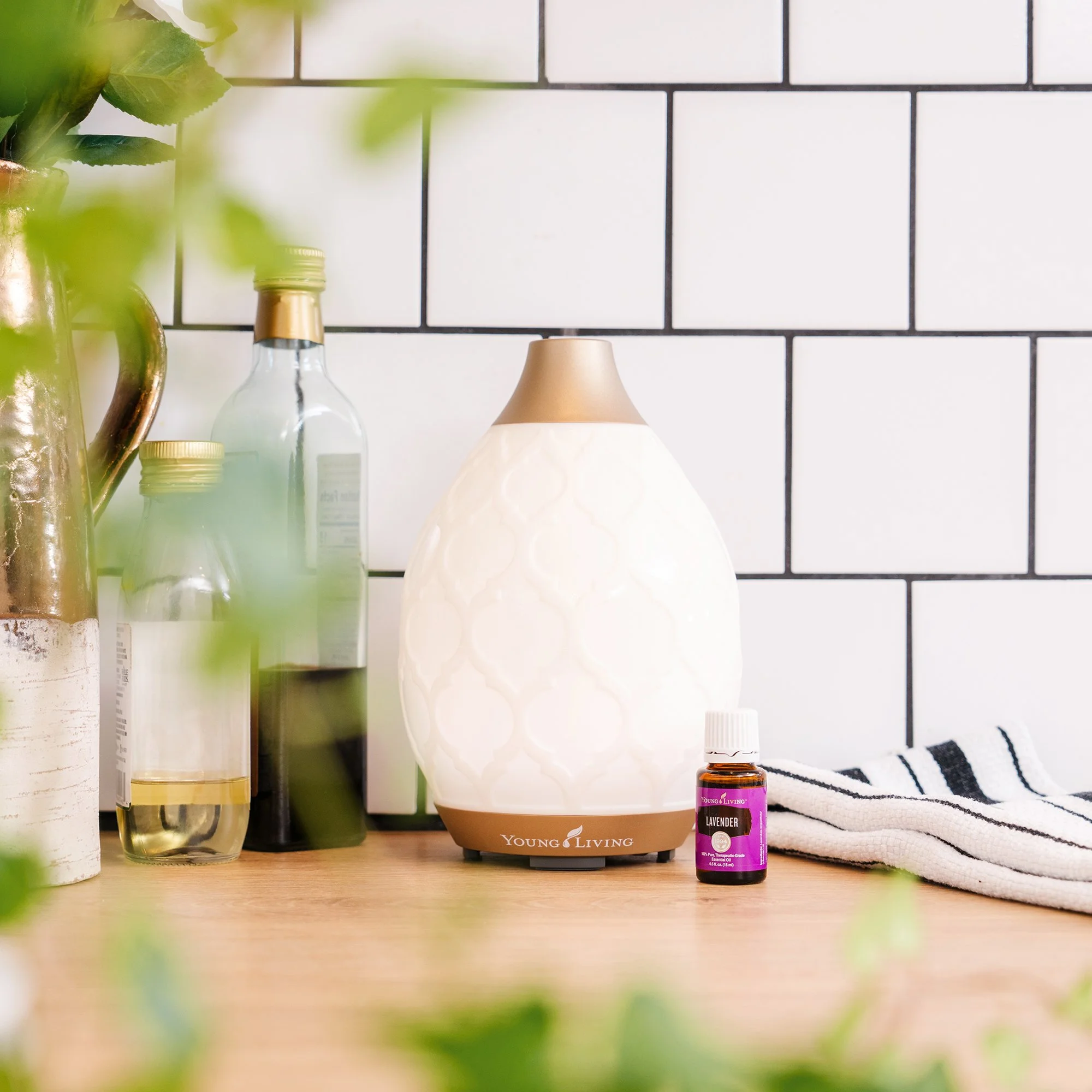 A Young Living lavender essential oil bottle beside a white aroma diffuser with a gold top, on a wooden surface with a striped cloth, against a white tiled wall.