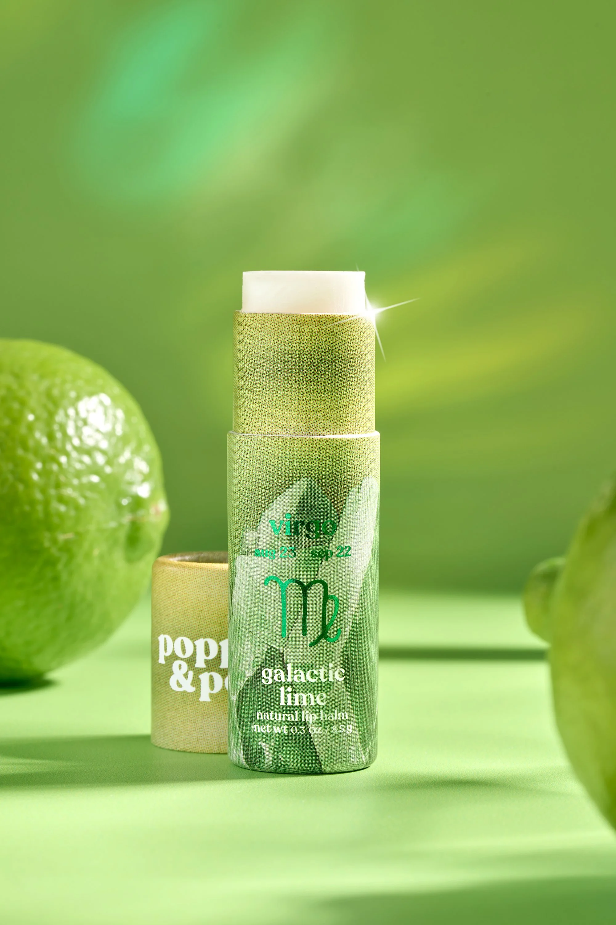 A green Poppy and Pout lip tube labeled 'galactic lime' with the zodiac sign Virgo and dates 'aug 23 - sep 22' in front of whole limes on a green background.