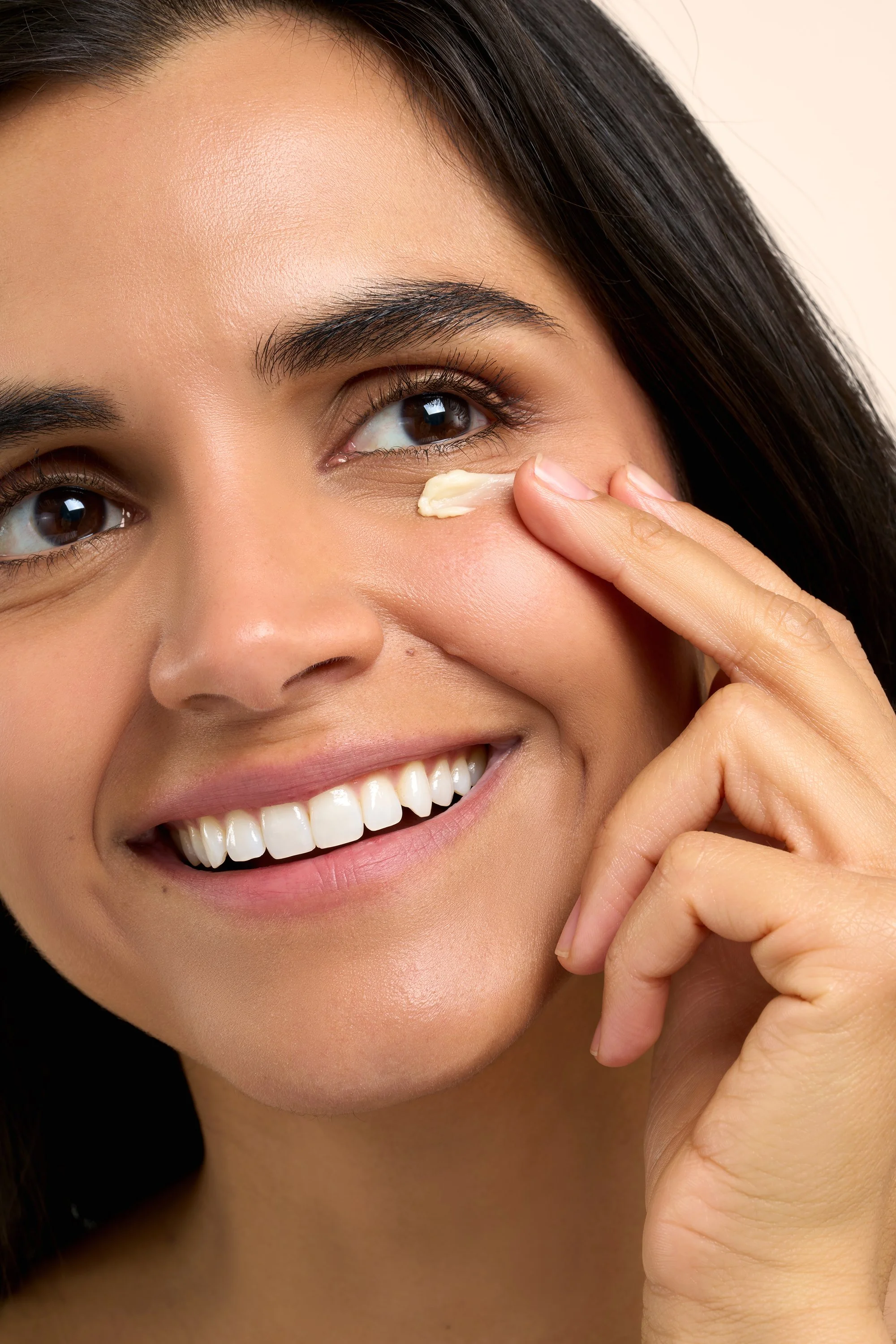 A woman smiling and applying a cream or moisturizer under her eye with her finger.