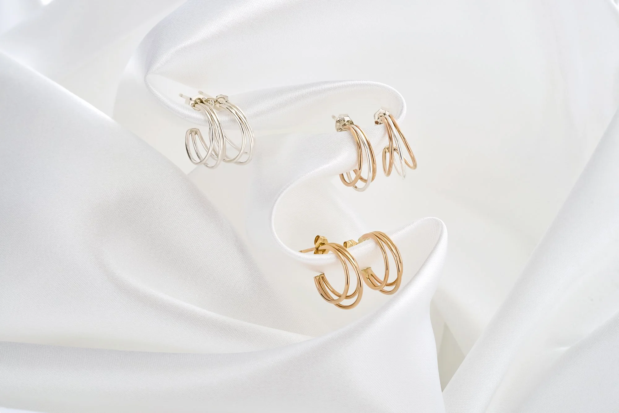 Three pairs of hoop earrings in gold and silver colors displayed on a white satin-like fabric.