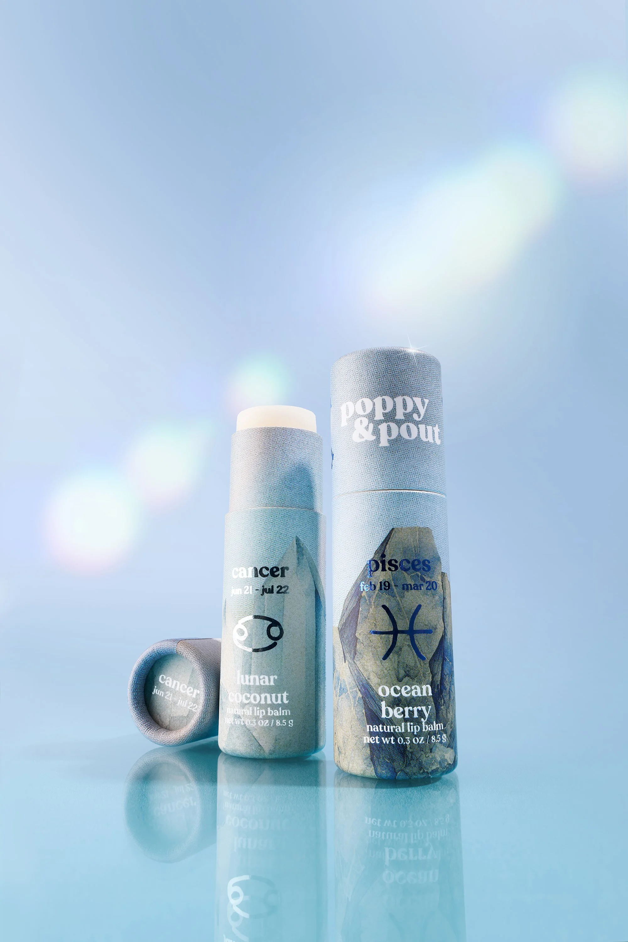 Two bottles of Poppy and Pout lip balm with zodiac signs and dates, one labeled Cancer and lunar coconut, the other labeled Pisces and ocean berry, against a blue background with reflections