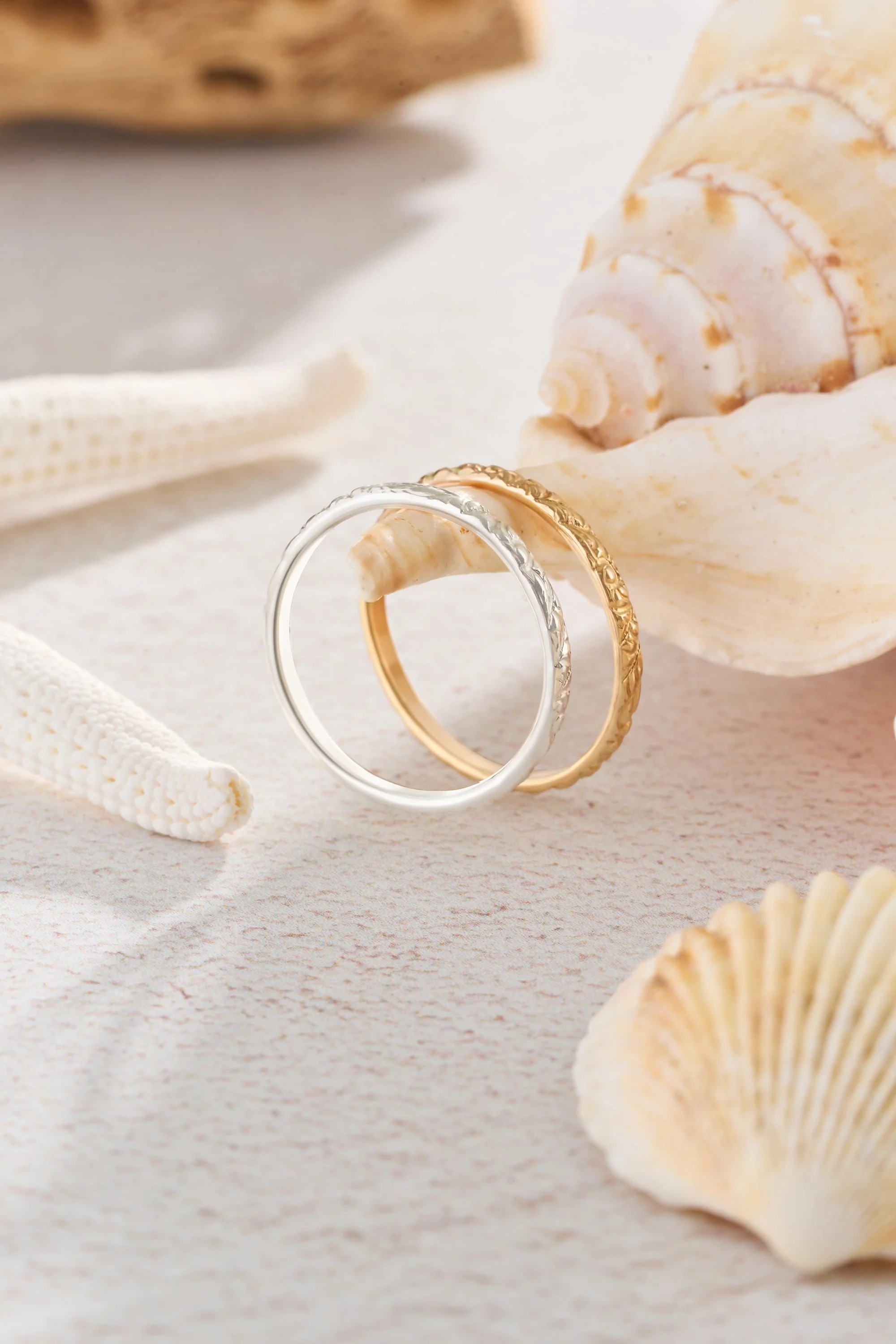 Two rings, one silver and one gold, resting on a seashell surrounded by additional seashells on a sandy surface.