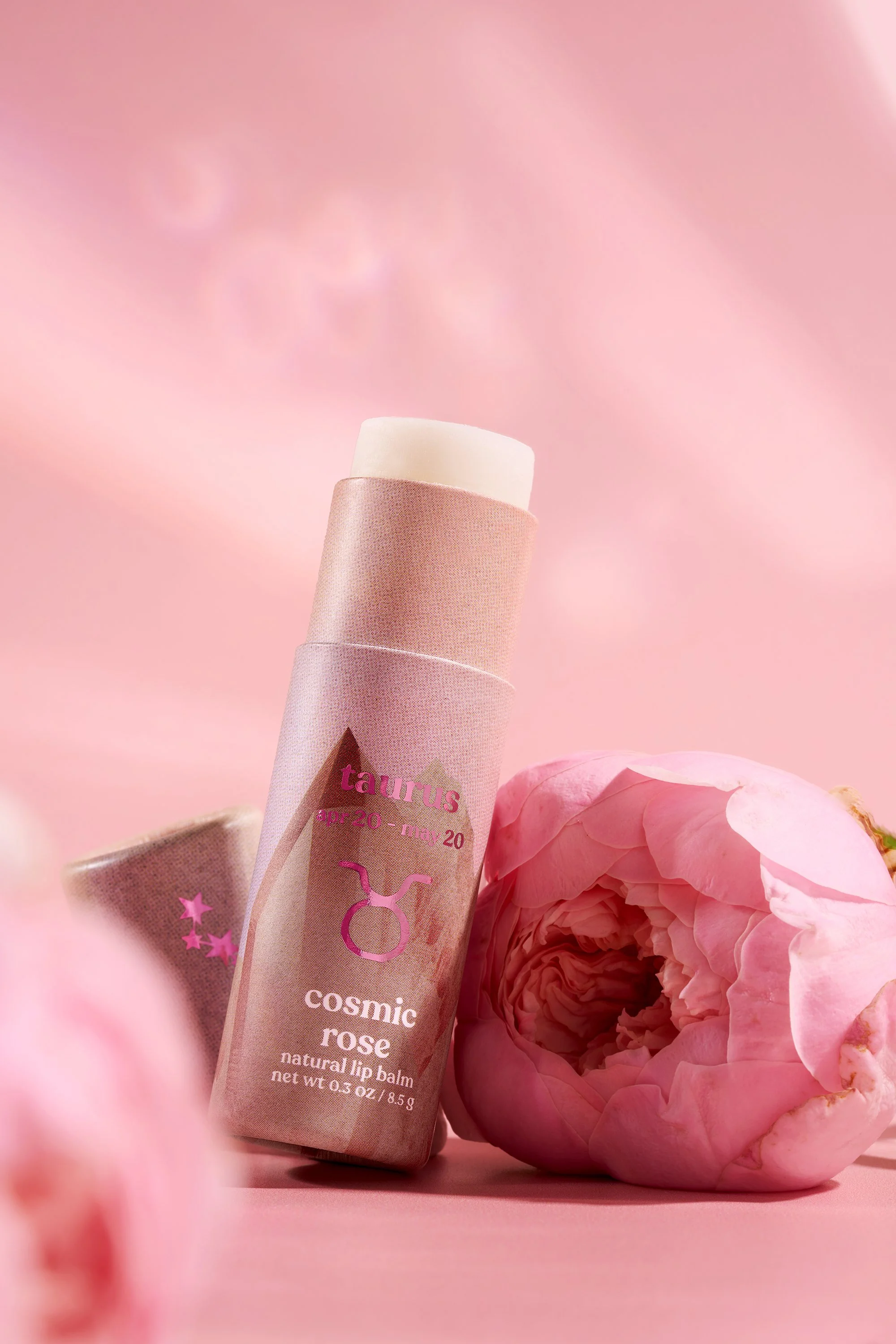A pink tinted tube of Poppy and Pout cosmic rose lip balm leaning against pink peonies on a pink background.