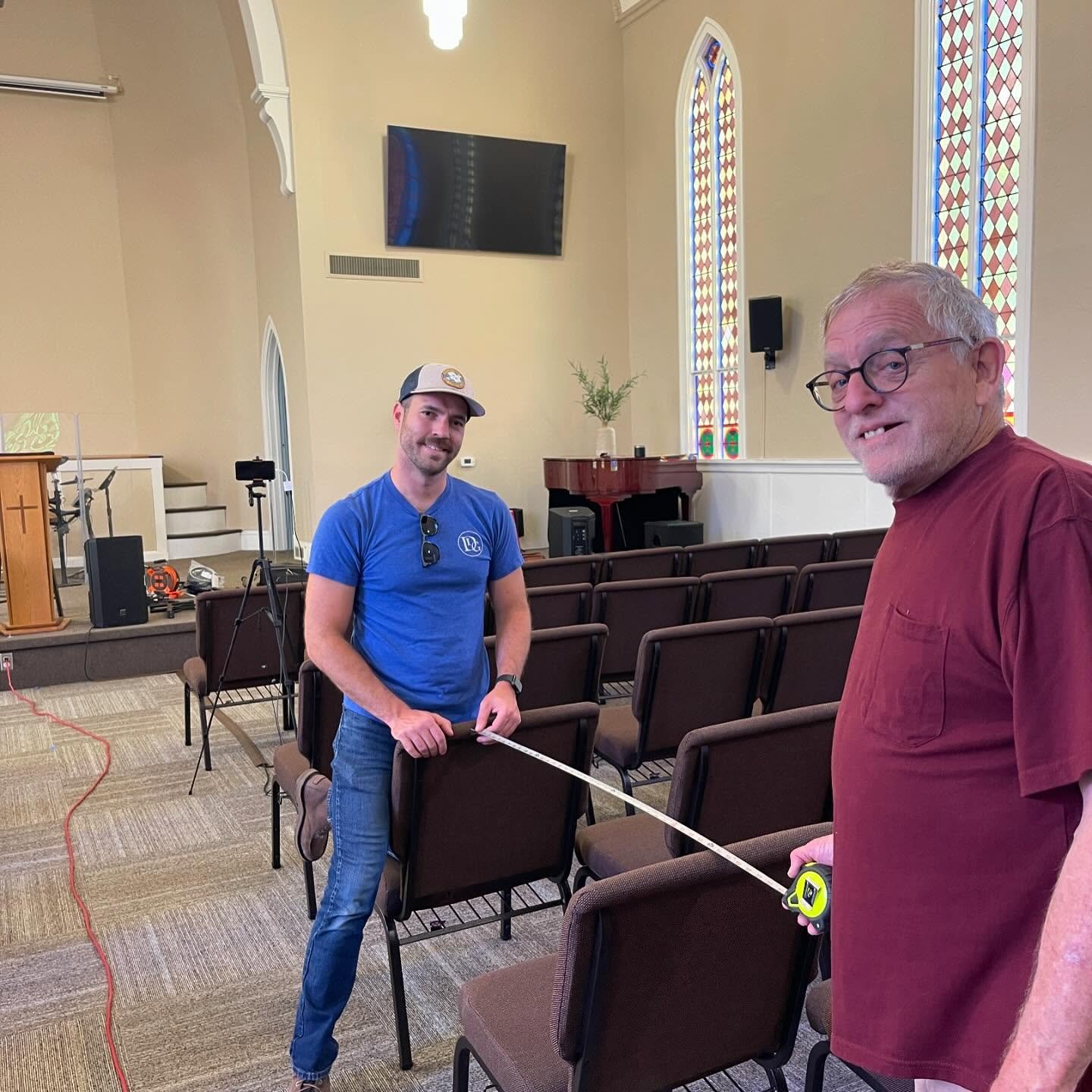 Two men standing in an empty church, measuring the distance between chairs with a tape measure, with rows of chairs, stained glass windows, a piano, and a large screen in the background.