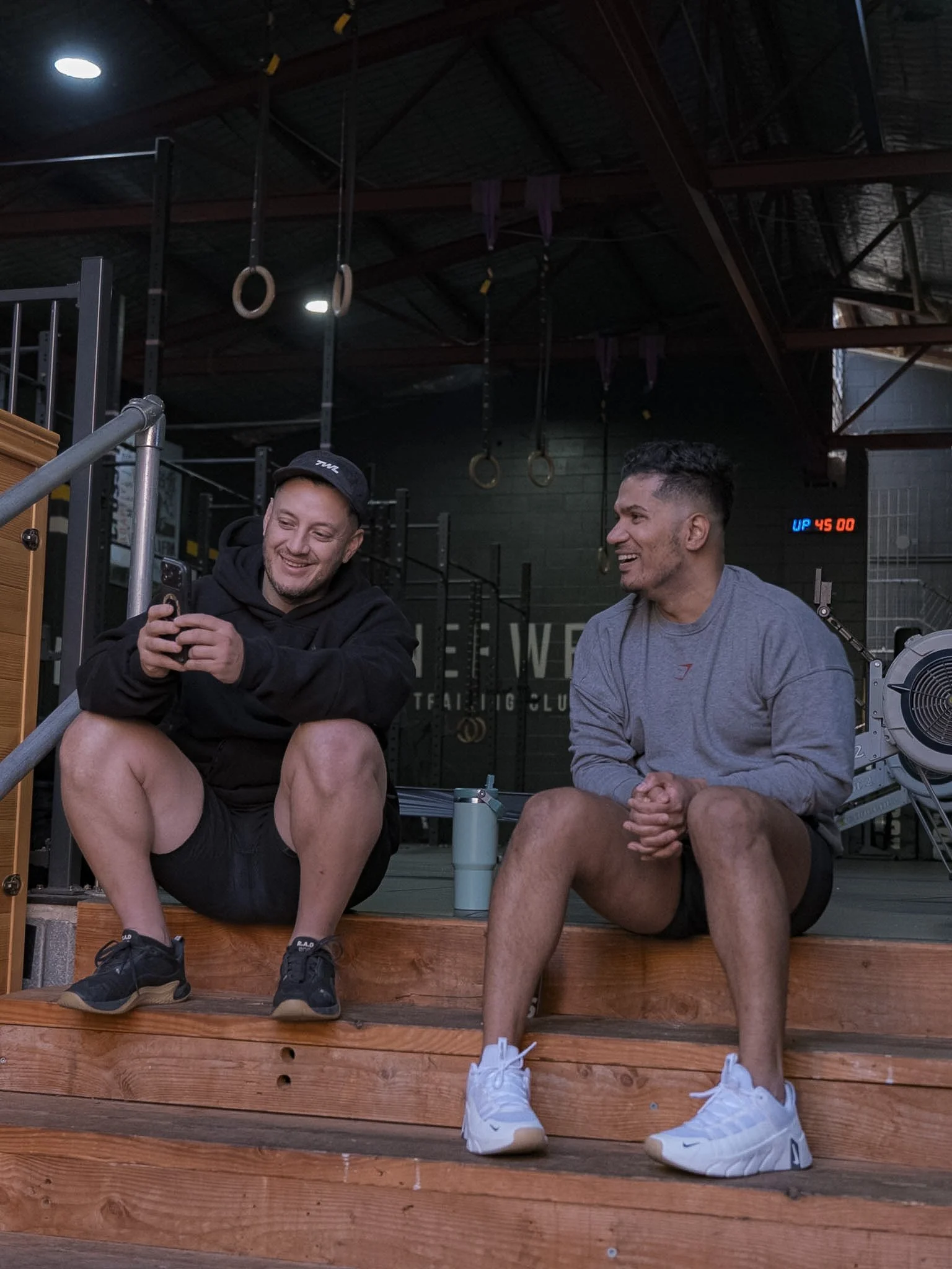 Two men sitting on a wooden bench in a gym, smiling and chatting. Gym equipment and hanging gymnastic rings are visible in the background.