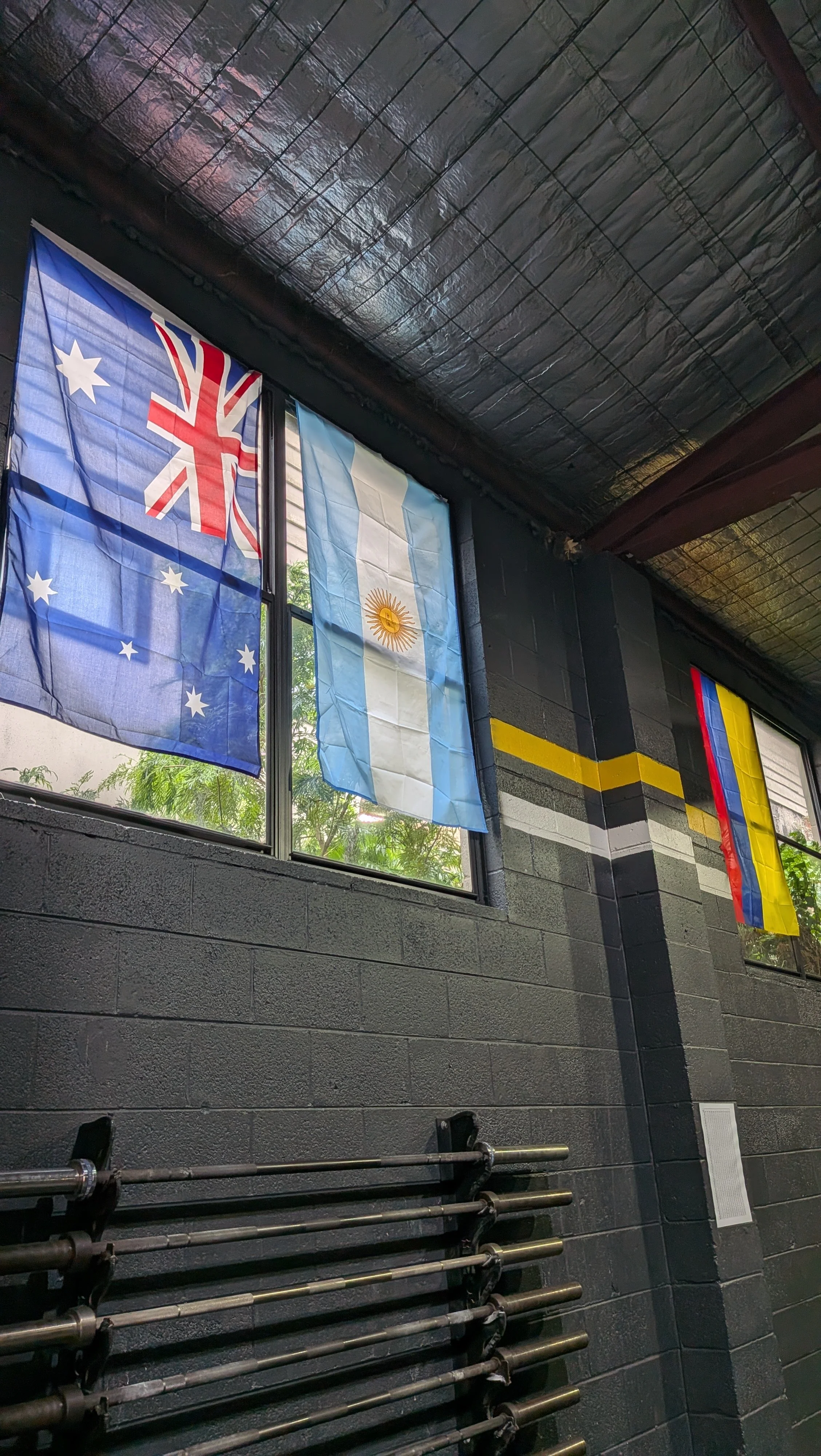 Indoor scene with three flags hanging in front of a window. The flags are of Australia, Argentina, and Colombia. The room has black brick walls and a black ceiling with some beams, with weights and a barbell on the floor beneath the flags.