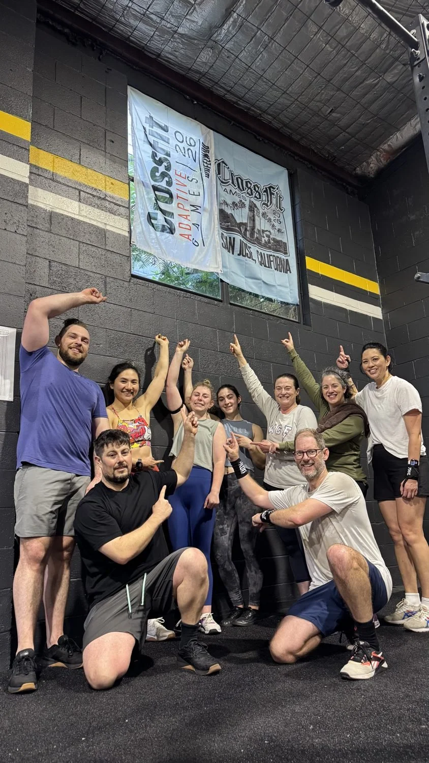 A group of nine people smiling and posing in a gym, pointing upward with their fingers. They are standing and kneeling in front of a black brick wall with banners hanging above them that say "CrossFit San Jose, California" and "CrossFit Adaptation Ga