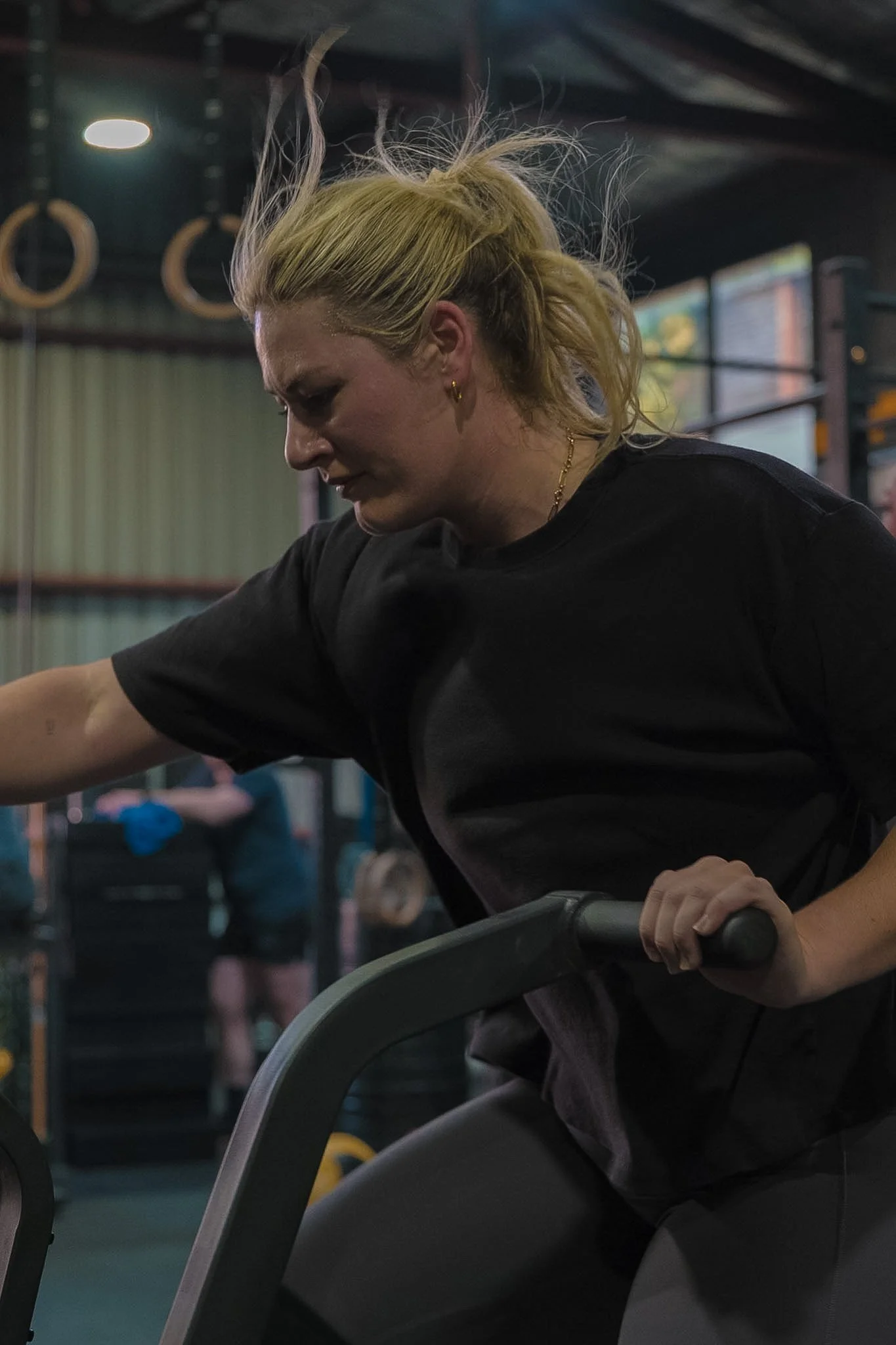 A woman with blonde hair tied back, wearing a black shirt, appears to be exerting effort while cycling indoors. The background shows gym equipment, including hanging gymnastic rings and a large window with curtains.