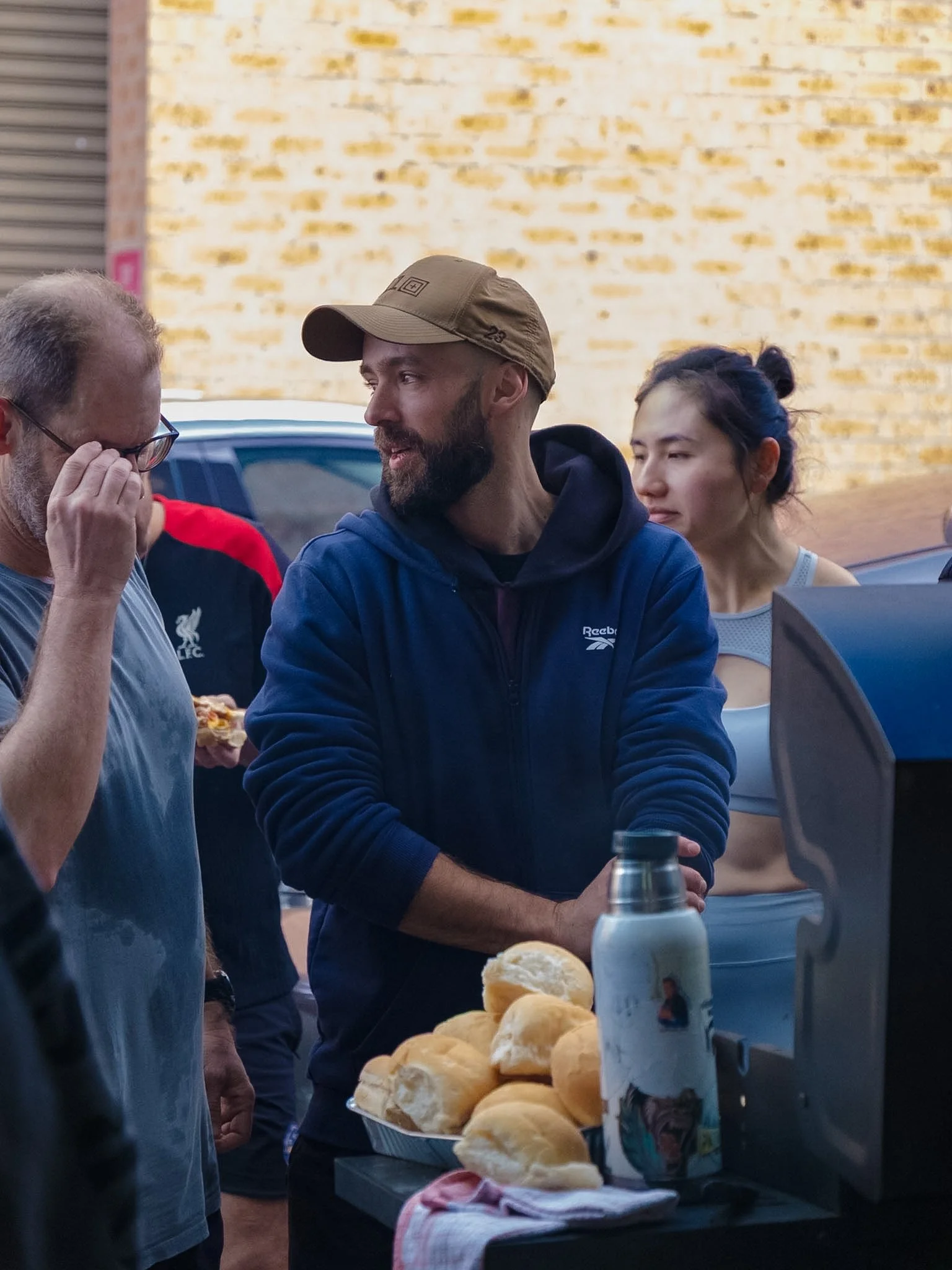 People at a bbq, with a tray of bread rolls and a bottle of water on the table, and a man in a blue hoodie talking with an older man.