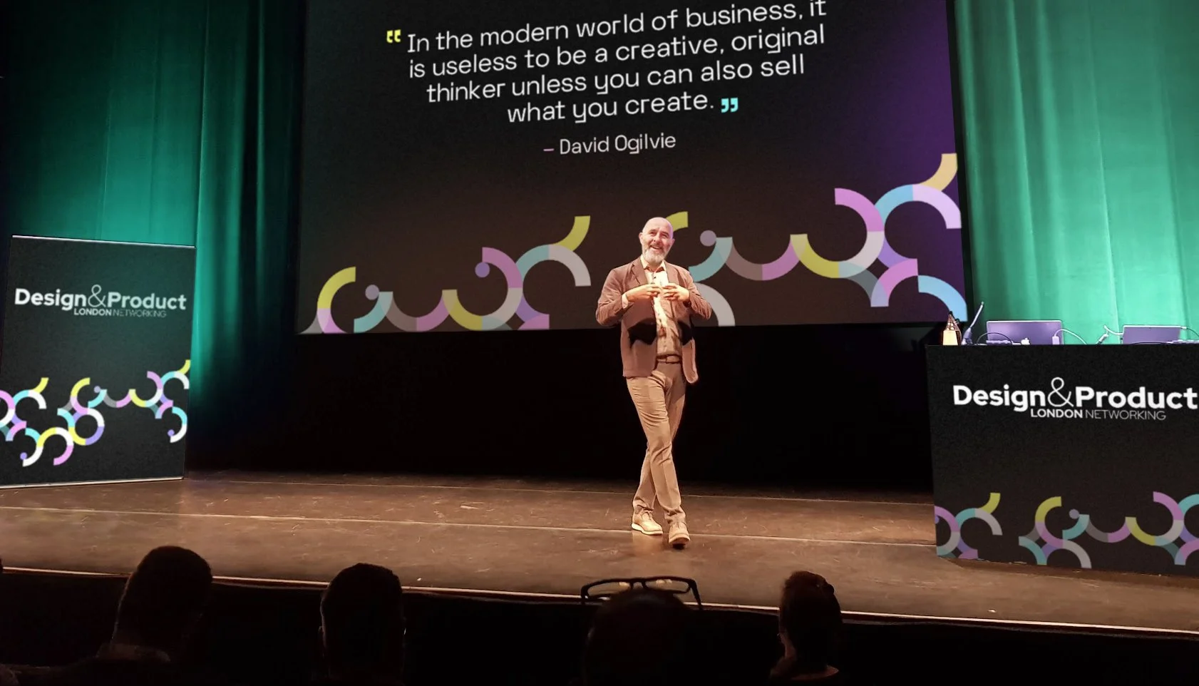 A speaker stands on a stage at the Design & Product London Networking event, with colorful abstract decoration and a large screen displaying a quote by David Ogilvie about creativity and business.