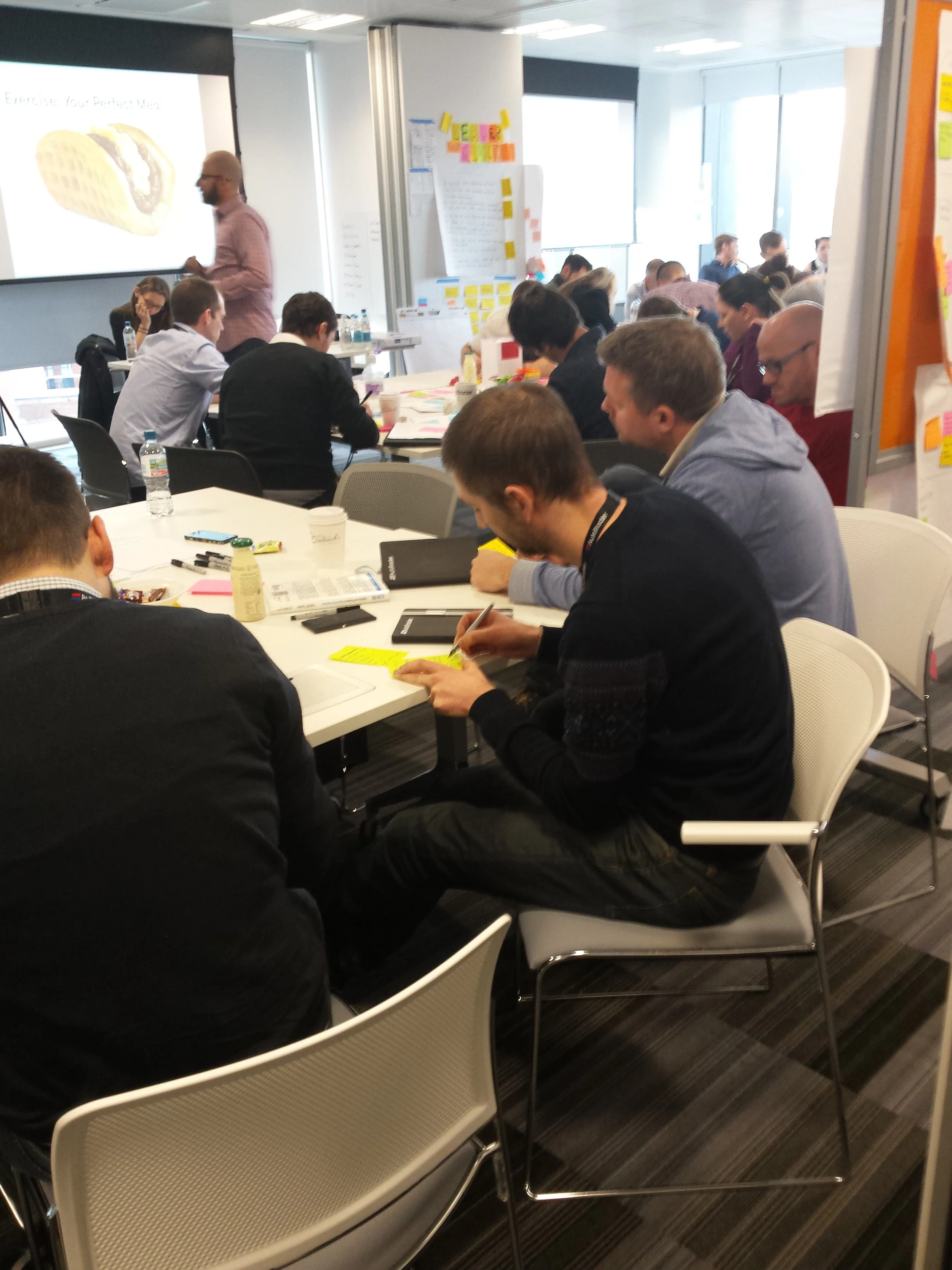 People attending a workshop or conference in a modern office setting, some taking notes, with a presenter at a screen displaying an image of a sandwich.