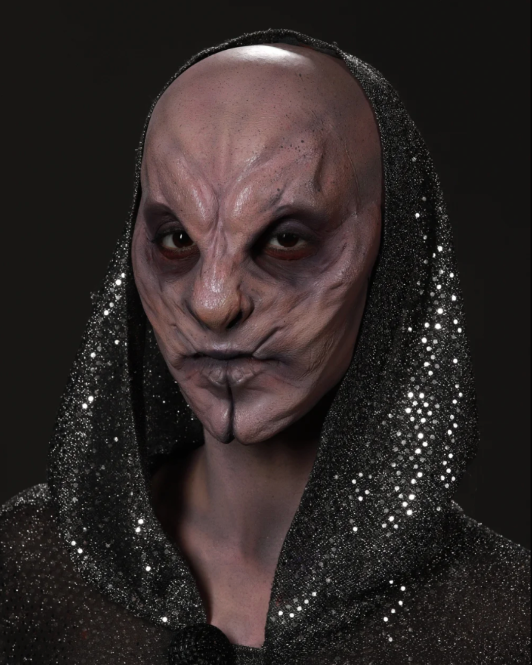 Person wearing a shiny black hood with a distorted, wrinkled face mask or makeup that resembles a creature or alien, with dark eye makeup, against a black background.
