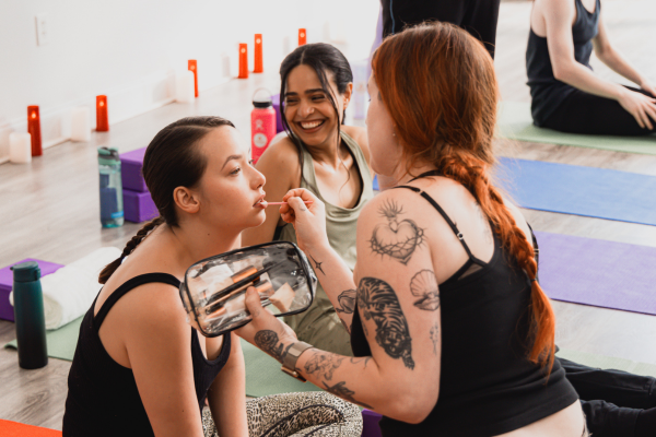 A woman with red hair and tattoos applies makeup or face paint to another woman with dark hair during a yoga or fitness class in a studio.