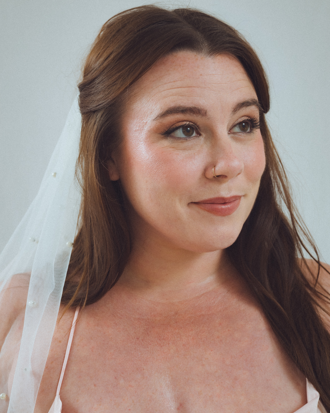 A close-up portrait of a woman with long brown hair, wearing a sheer white veil with small pearls, and a subtle makeup look, with a neutral background.