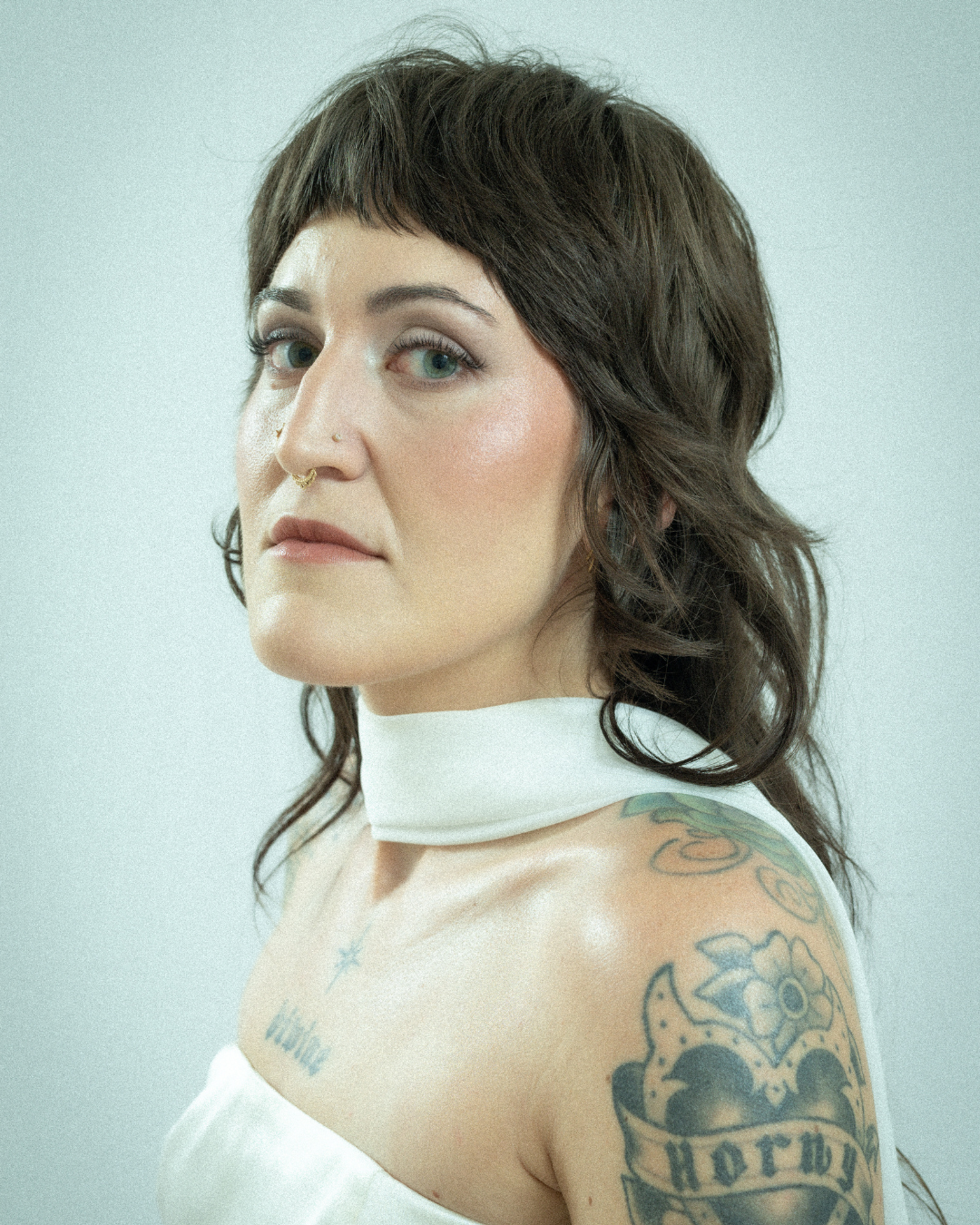 Portrait of a woman with dark brown hair, tattoos on her shoulder and chest, wearing a white top and a white choker, looking at the camera with a neutral expression.