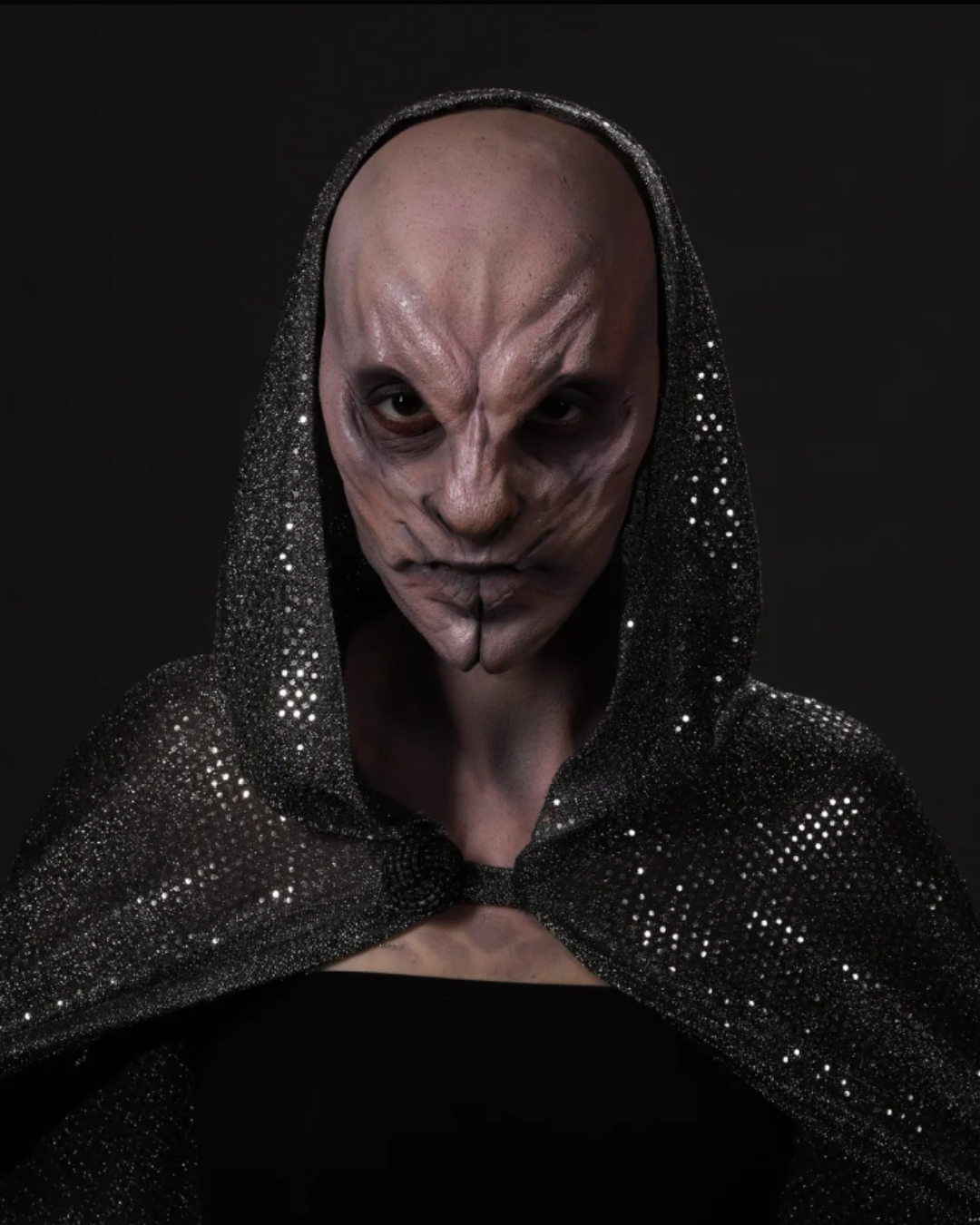 Person with a bald head and elaborate face makeup resembling a creature or alien, wearing a black, sparkly hooded cloak.