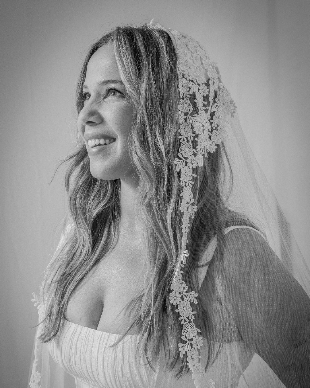 Black and white portrait of a smiling woman wearing a wedding veil with lace details.