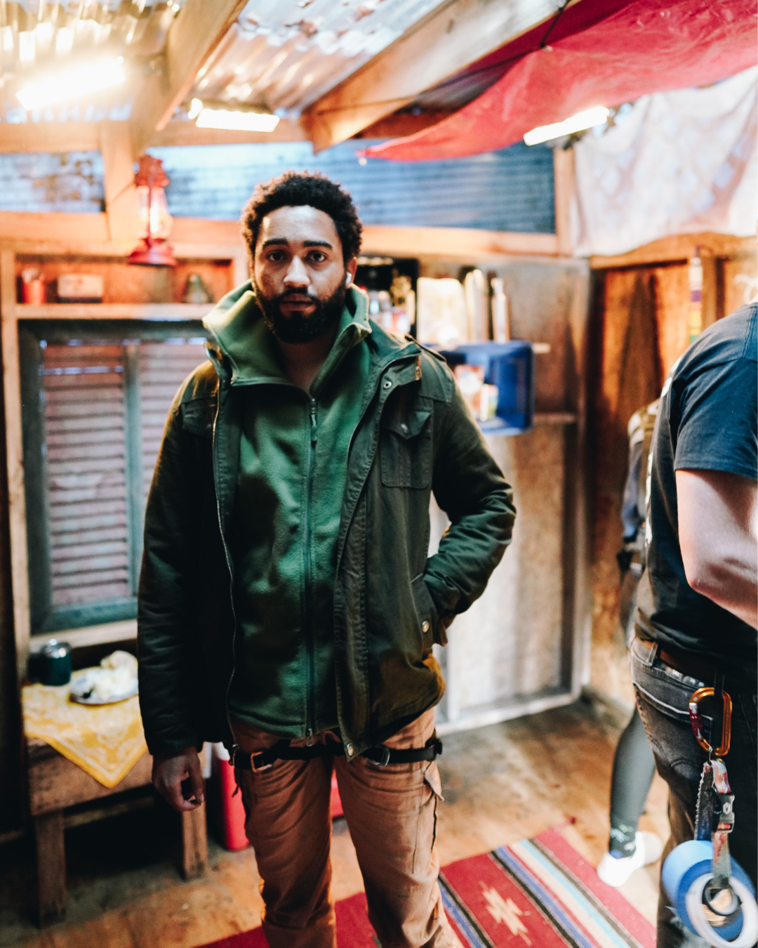 A man with a beard and curly hair, wearing a dark green jacket over a green hoodie, standing inside a rustic, wood-paneled room with warm lighting.