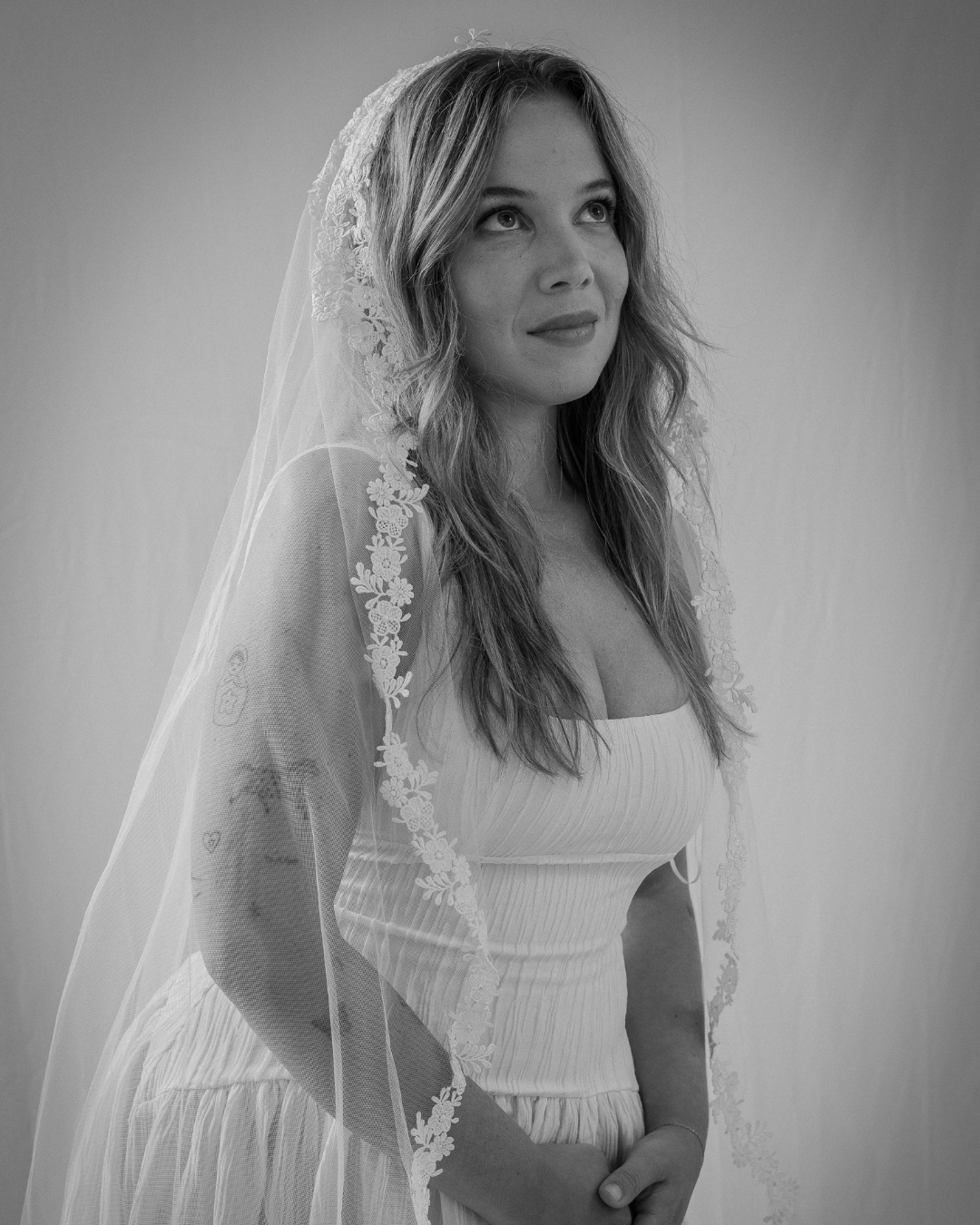 Black and white photo of a woman wearing a wedding dress and veil, looking upward with a gentle smile.